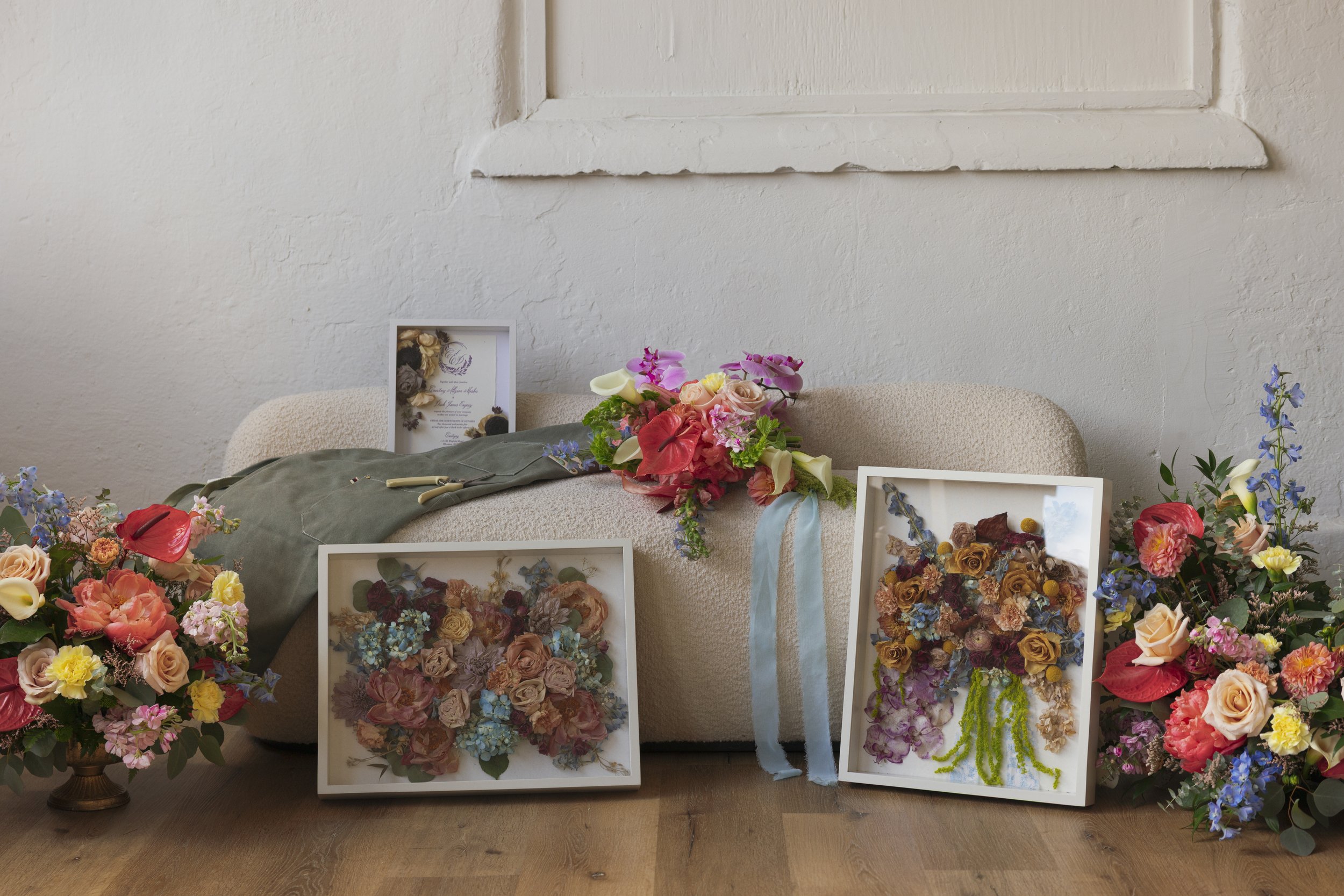 Arranged floral displays and framed pressed flower art on a beige sofa and wooden floor.