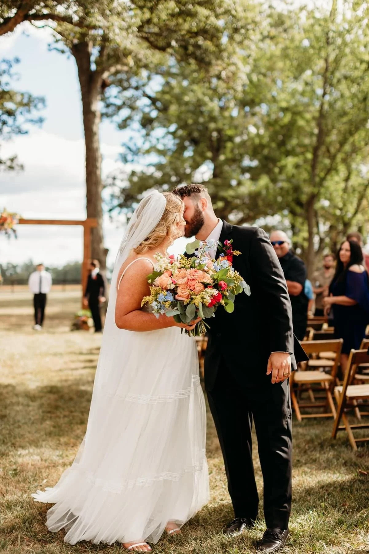 A bride and groom share a kiss at their outdoor wedding ceremony, with the bride holding a colorful bouquet and guests watching in the background.