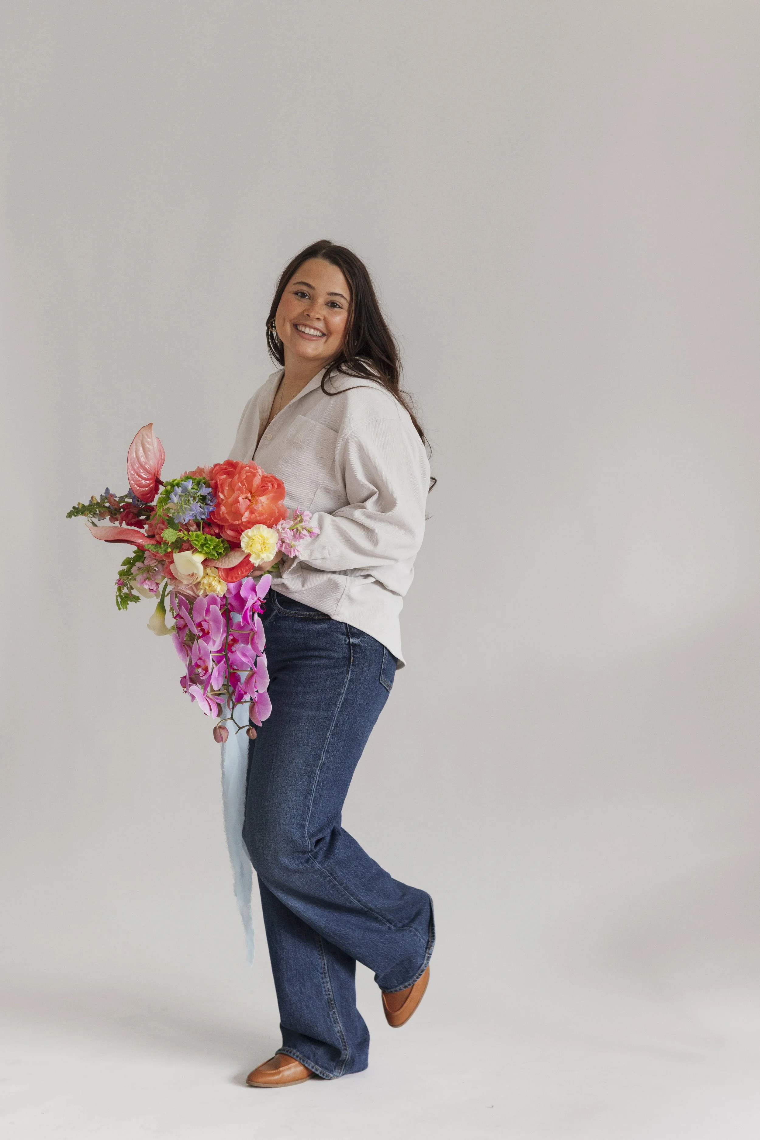 A woman stands holding a large bouquet of colorful flowers and smiling at the camera, wearing a beige top and blue jeans, against a plain white background.