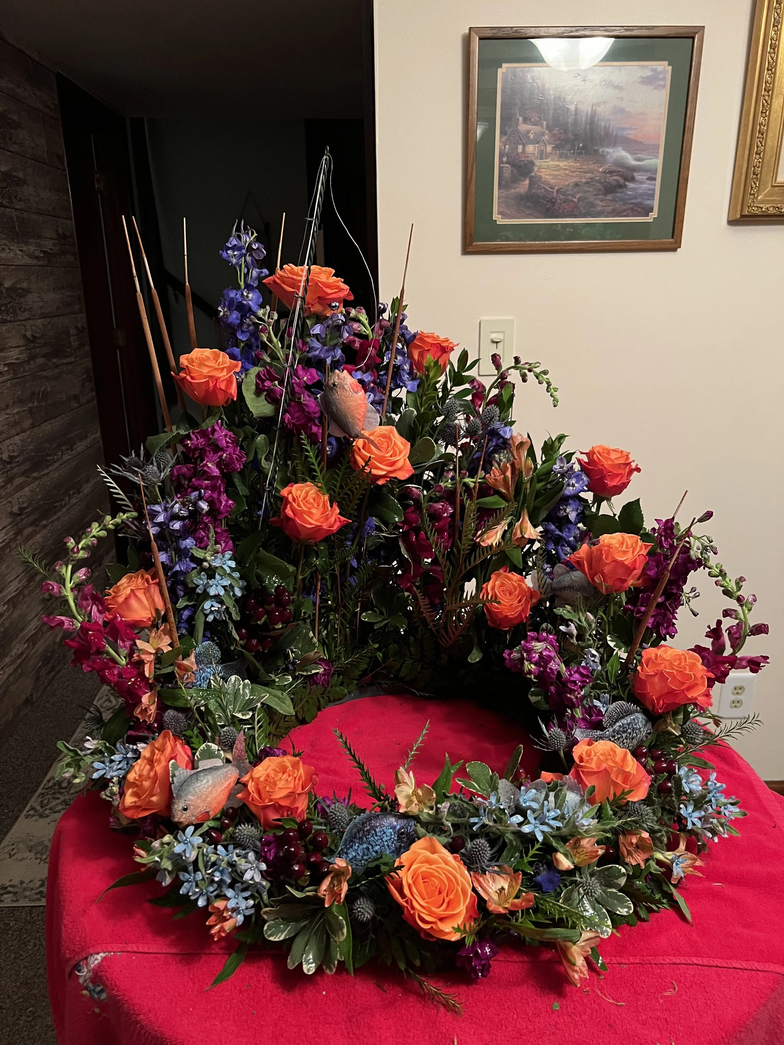 Colorful floral arrangement with orange roses, purple and blue flowers, green foliage, decorated with bird and feather accents, placed on a red cloth.