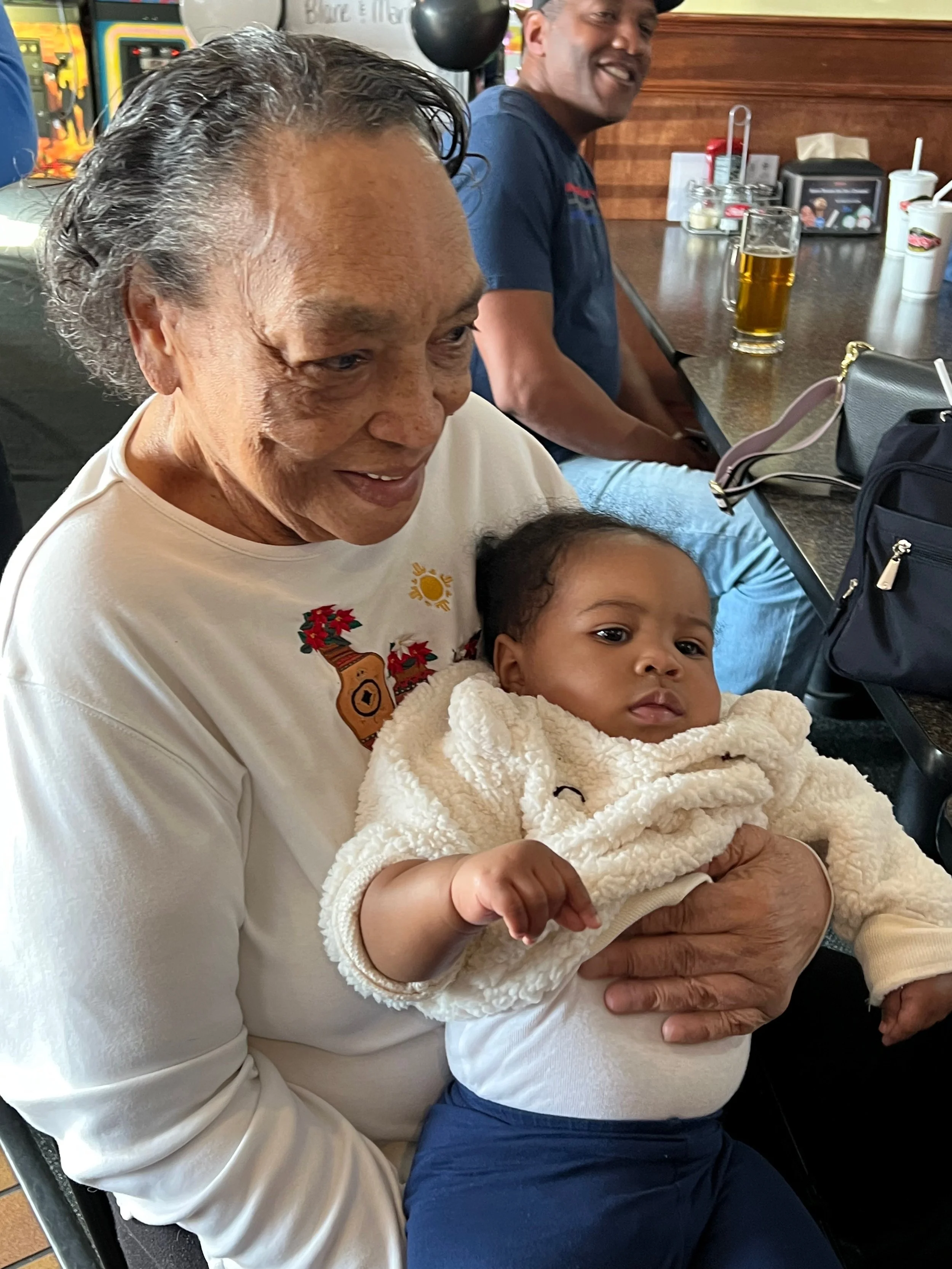 Older woman holding a baby in a restaurant or cafe. The woman is smiling, and the baby is looking away with a neutral expression. In the background, a man is sitting at the counter with a glass of beer, and various condiments and items are on the cou
