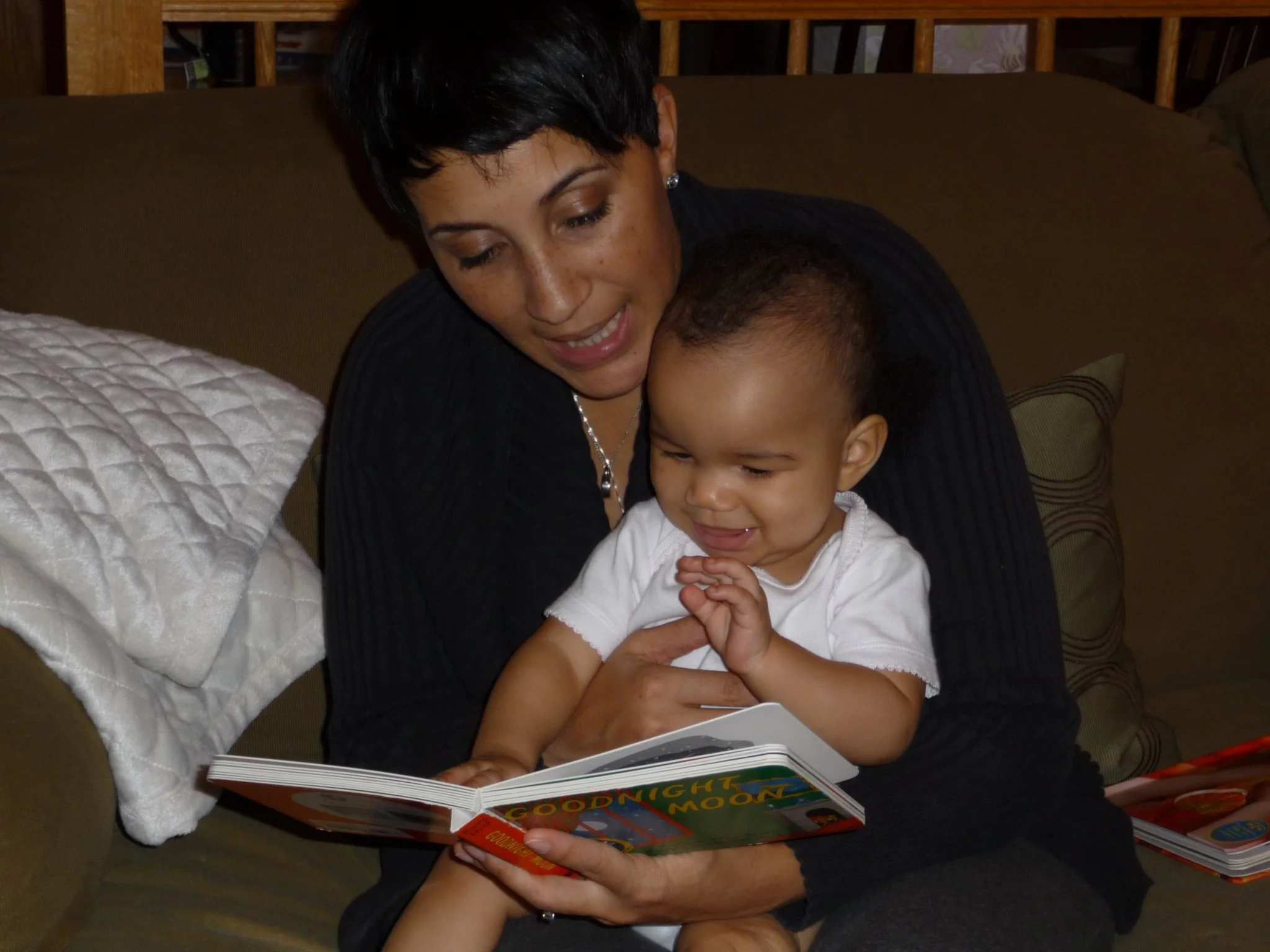 A woman with short dark hair and a boy with short curly hair sitting on a brown couch, smiling and reading a colorful children's book titled 'Goodnight Moon'.