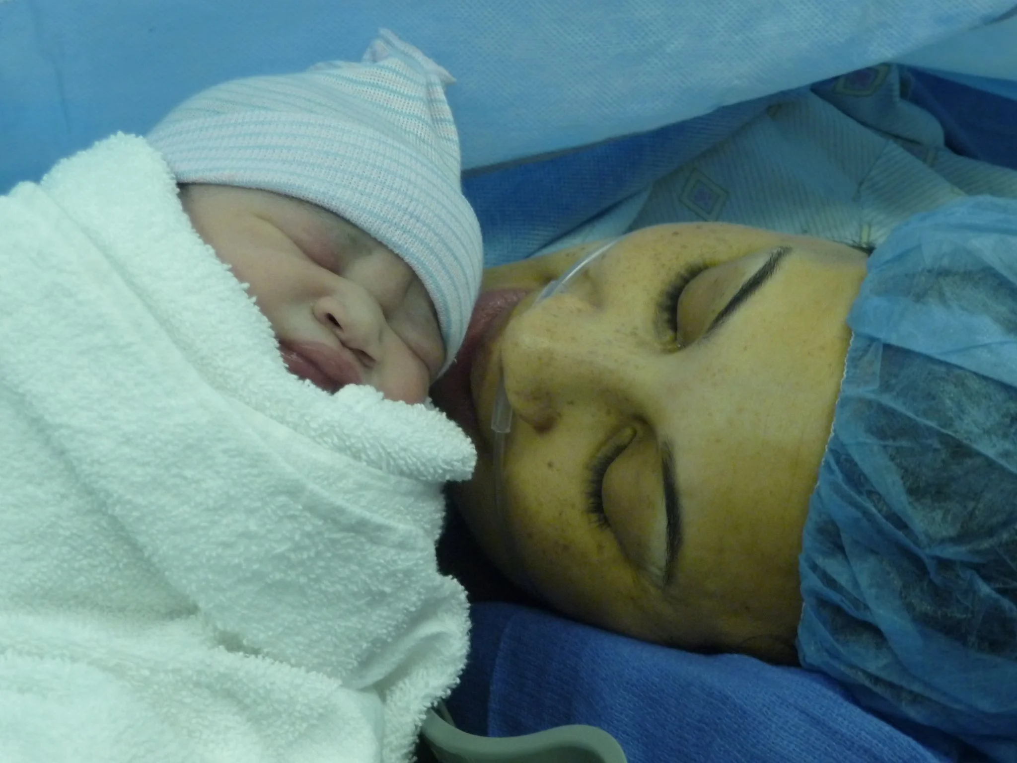 A newborn baby sleeping on a woman's chest in a hospital bed. The woman is wearing a blue surgical cap, and the baby is dressed in a knit cap and a white blanket.