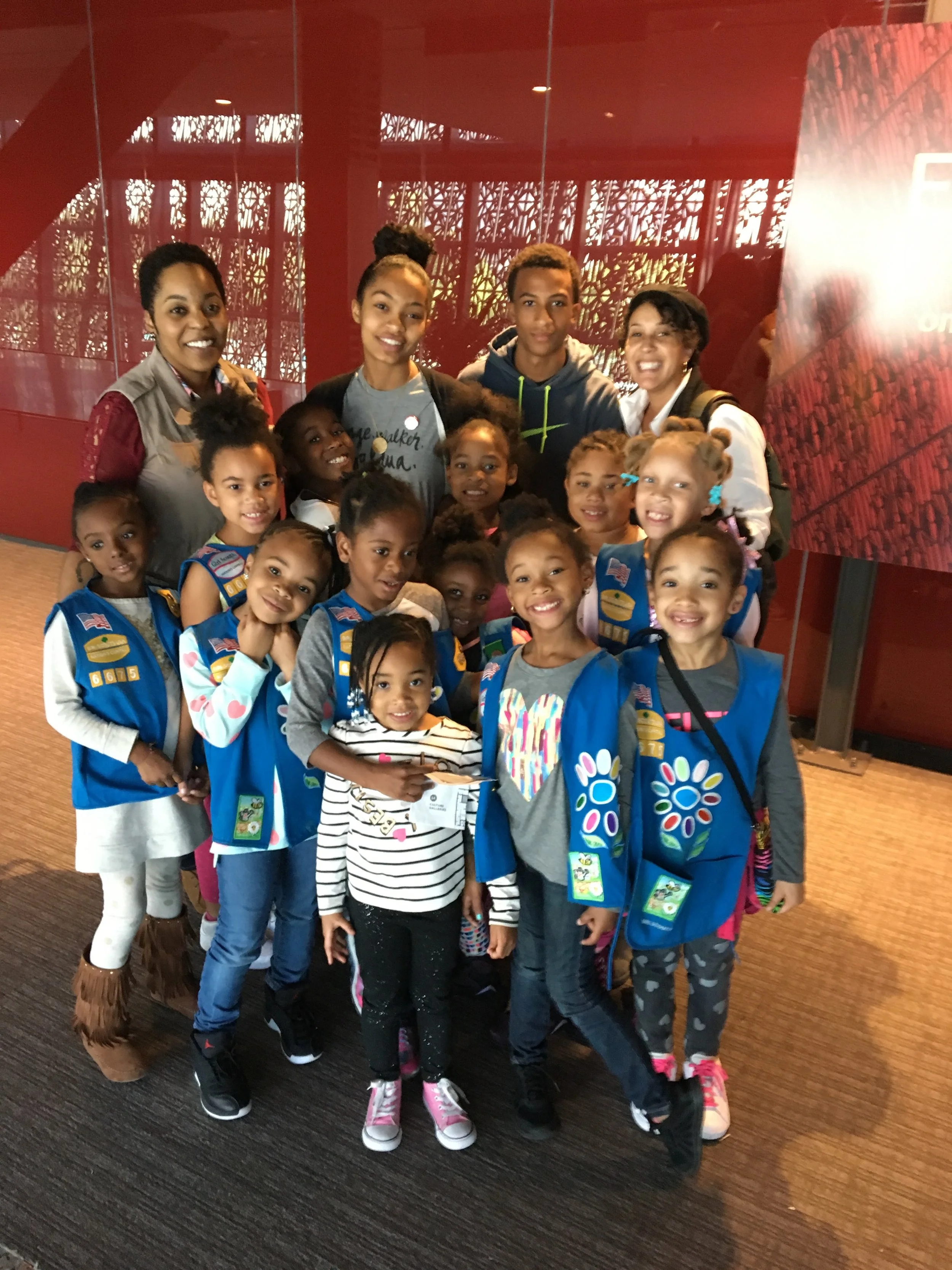 Group of children and adults posing together indoors, some children wearing blue scout vests with patches.