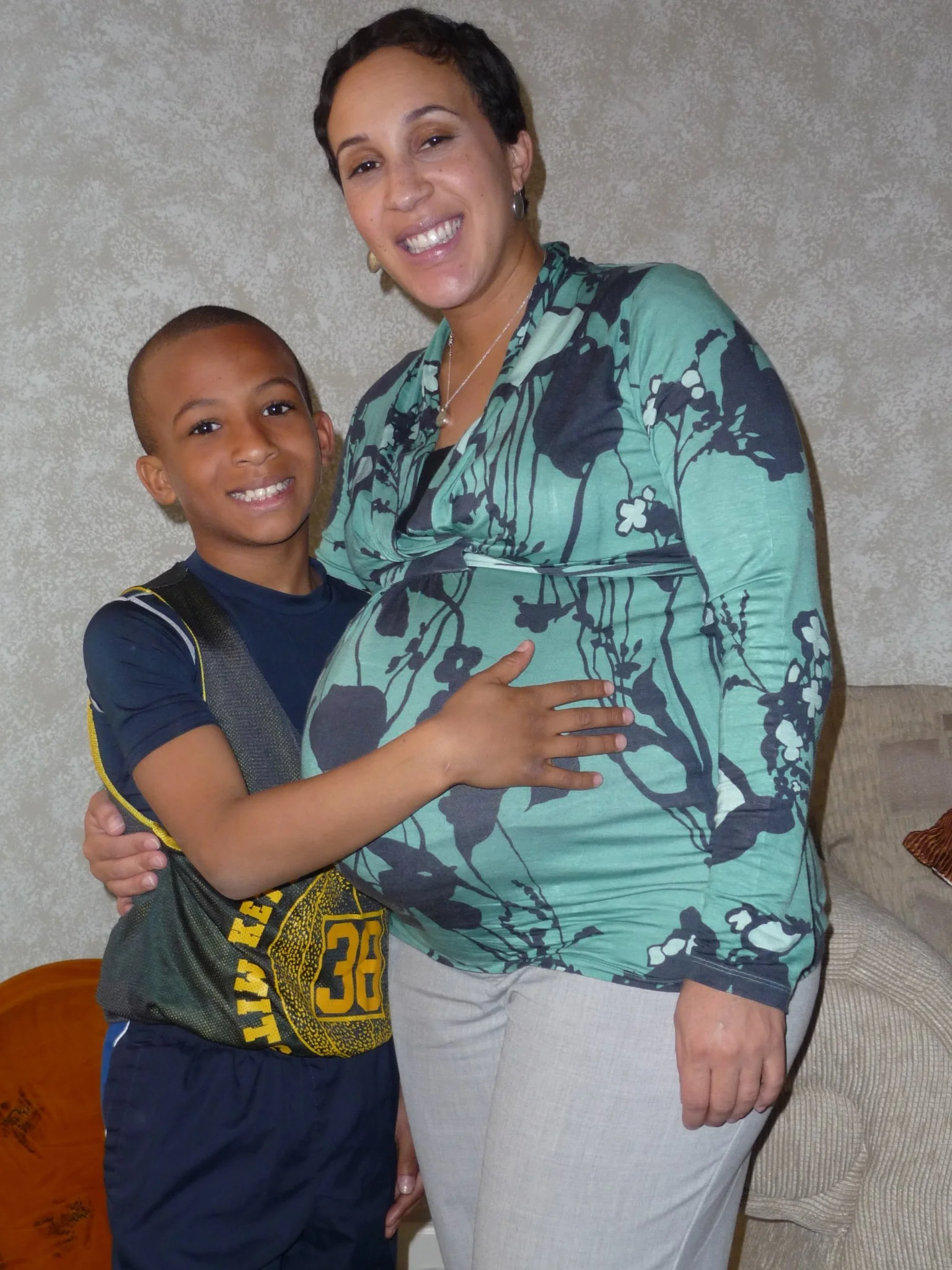 A pregnant woman smiling while hugging a young boy who is also smiling, standing indoors.