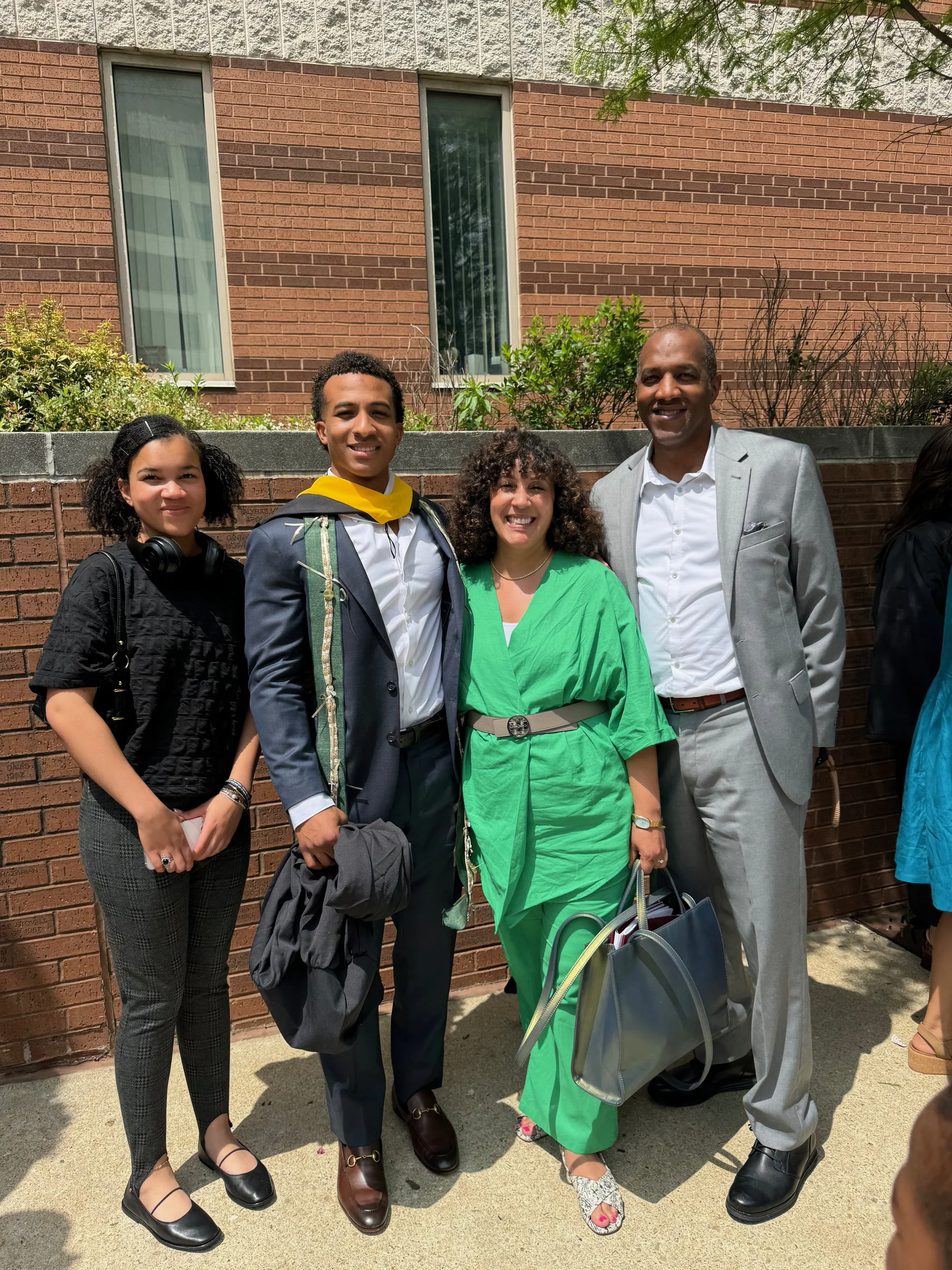 Group of five people standing outdoors in front of a brick wall, dressed in formal and semi-formal attire, smiling for a photo.