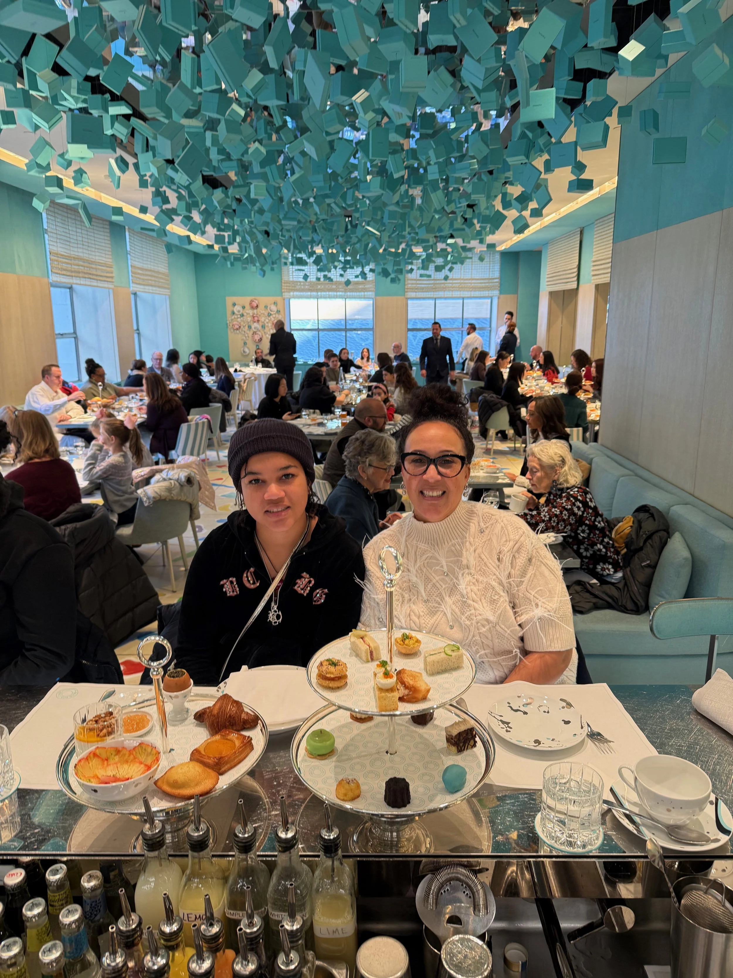 A woman and a girl enjoying high tea at a restaurant with an elegant atmosphere, featuring assorted sandwiches, desserts, and beverages on a tiered tray in front of them, with many other diners in the background.