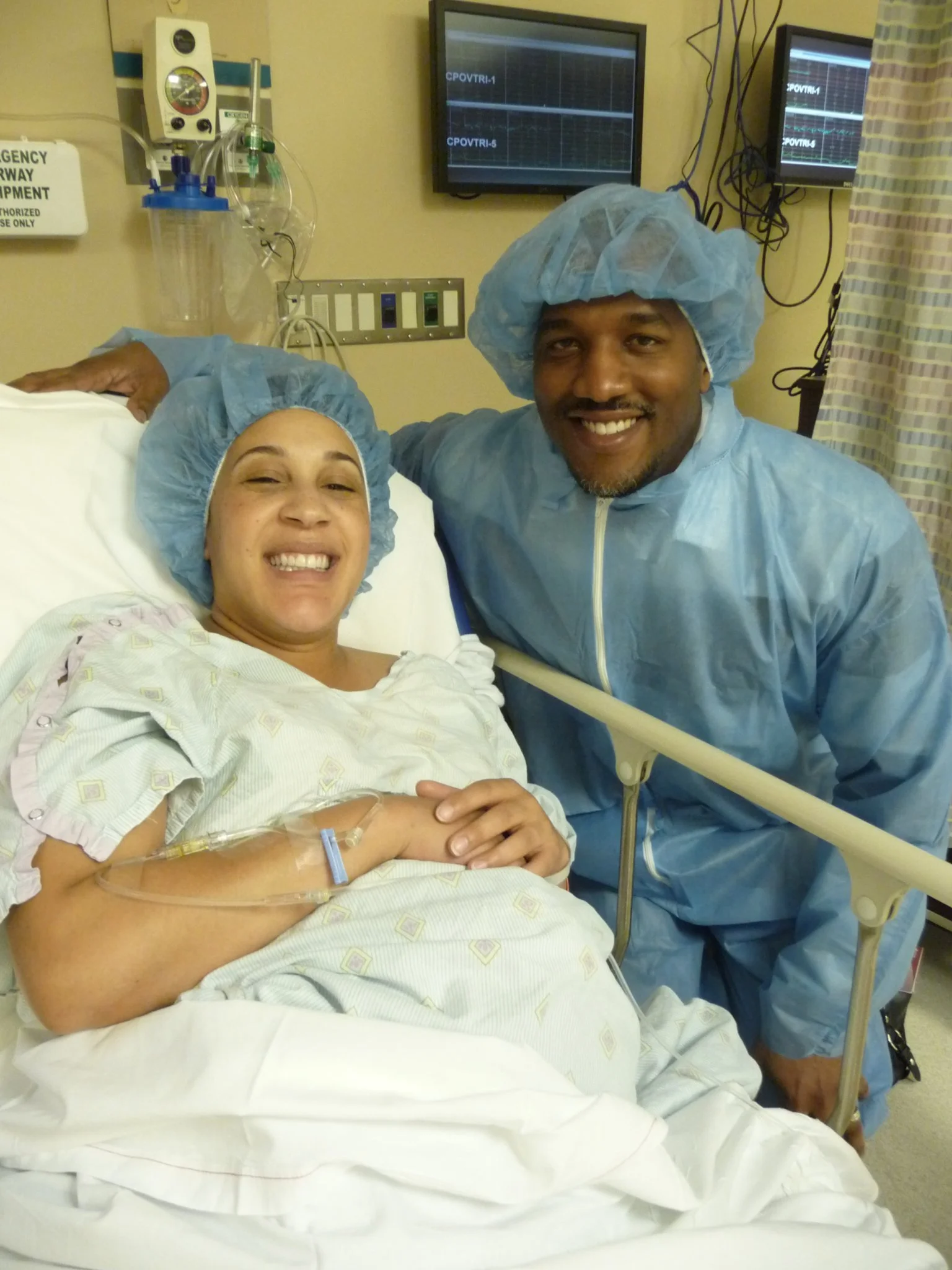 A woman in a hospital bed smiling, with a man in hospital gown and scrubs standing beside her. Both are wearing blue medical caps.