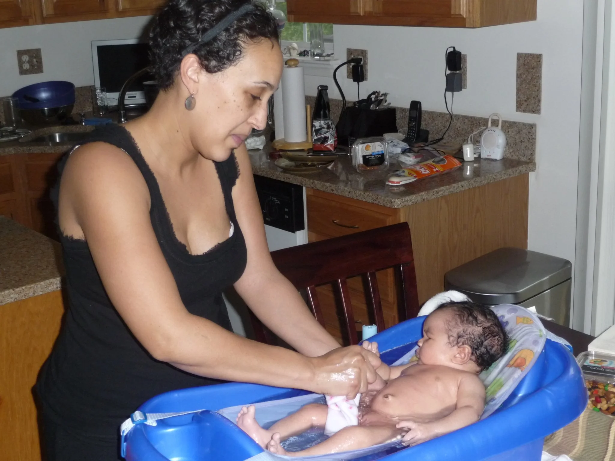 A woman giving a bath to a baby in a blue plastic infant bathtub in a kitchen.