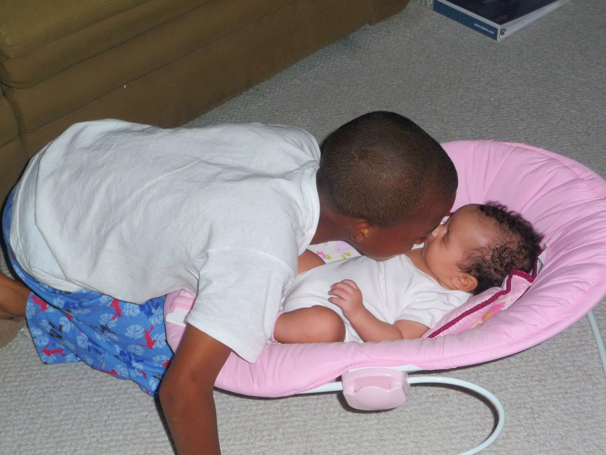 A young boy leaning over to kiss a baby girl who is lying in a pink baby bouncer on a carpeted floor.