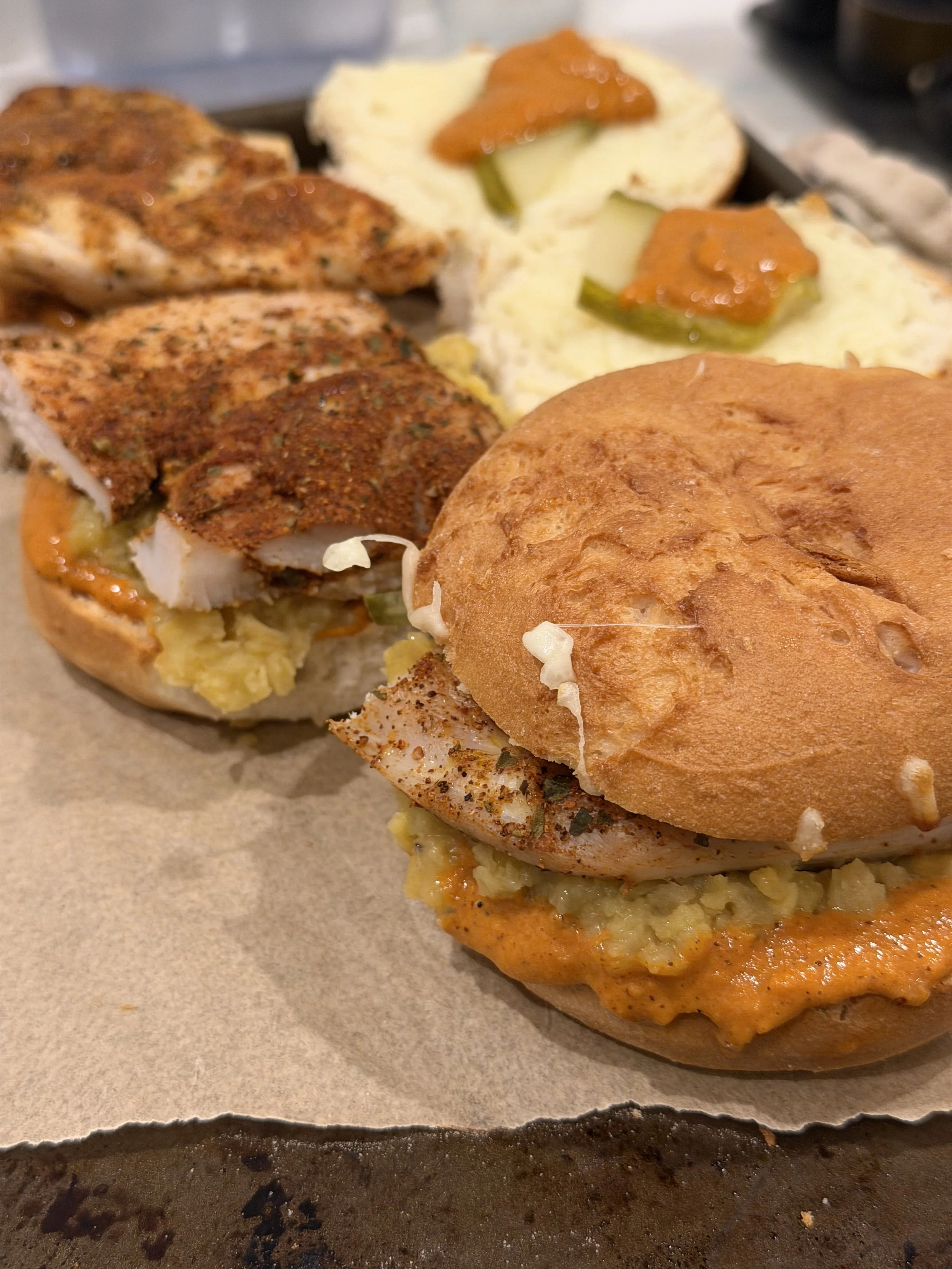 Two chicken sandwich sliders with seasoned chicken, mayonnaise or sauce, lettuce, and pickle slices, served on toasted buns, with mashed potatoes topped with gravy in the background.