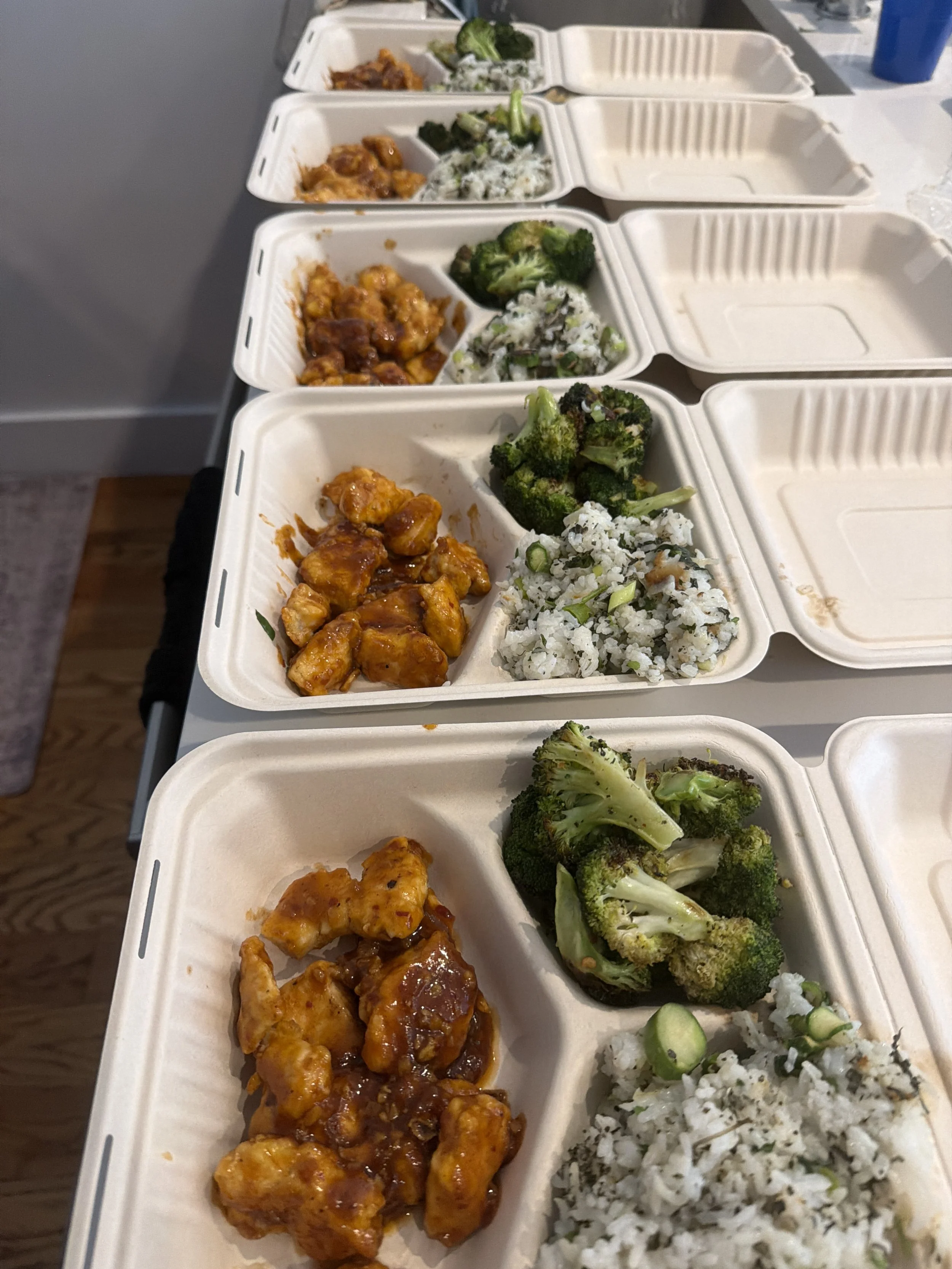 Multiple takeout containers filled with various Asian dishes, including spicy chicken, broccoli, and rice. The containers are aligned on a table.