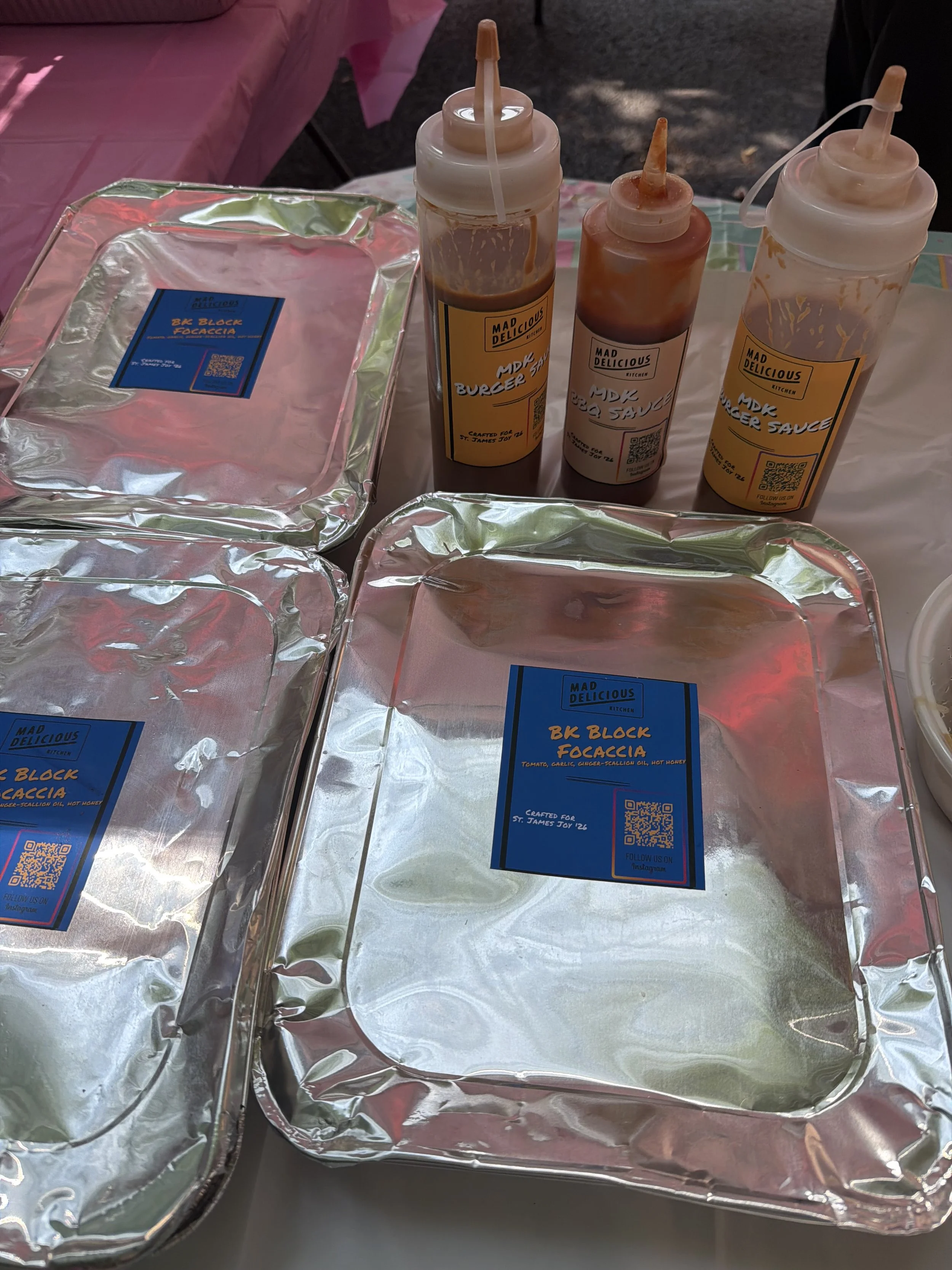 Three squeeze bottles of burger sauce on a tray, with foil food containers and pink tablecloths in the background.