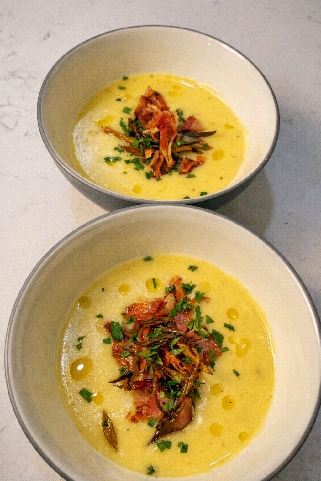 Two bowls of creamy yellow soup topped with chopped herbs, crispy bacon, and shredded vegetables, drizzled with oil.