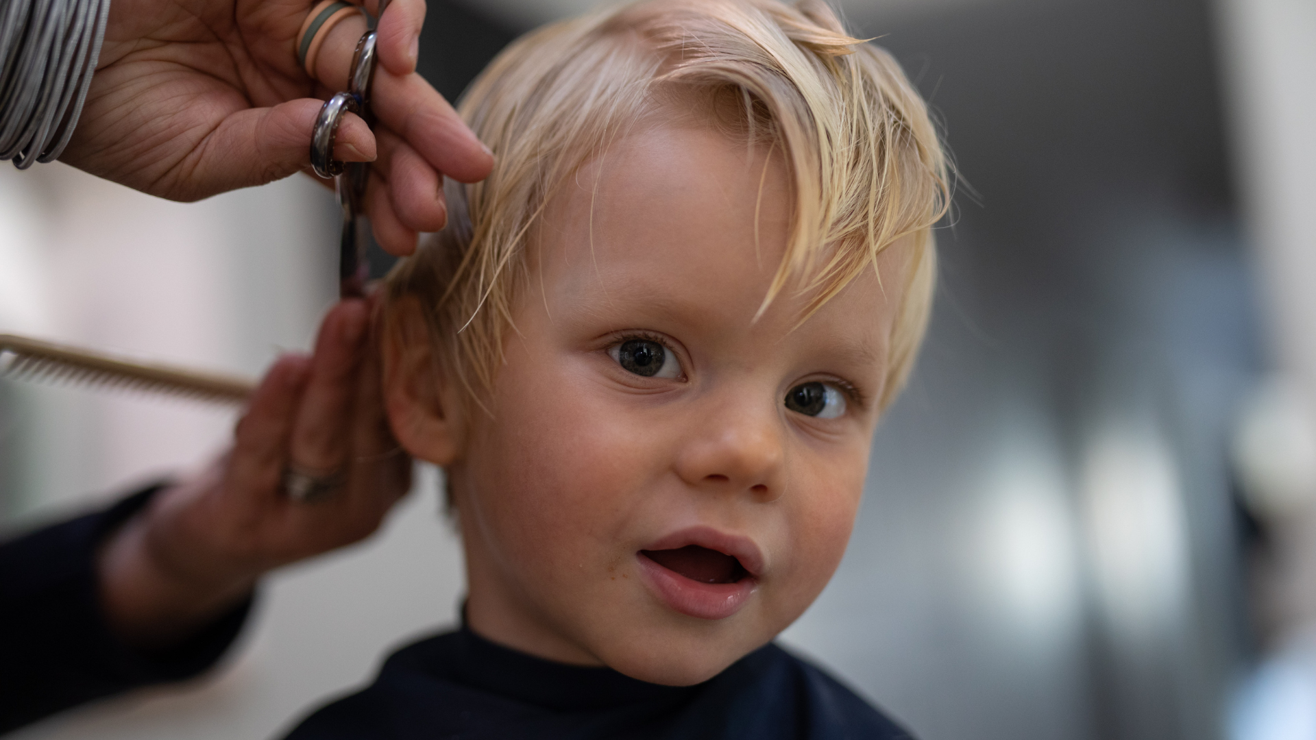 A young boy with blond hair getting a haircut at a salon, looking at the camera with an open mouth.