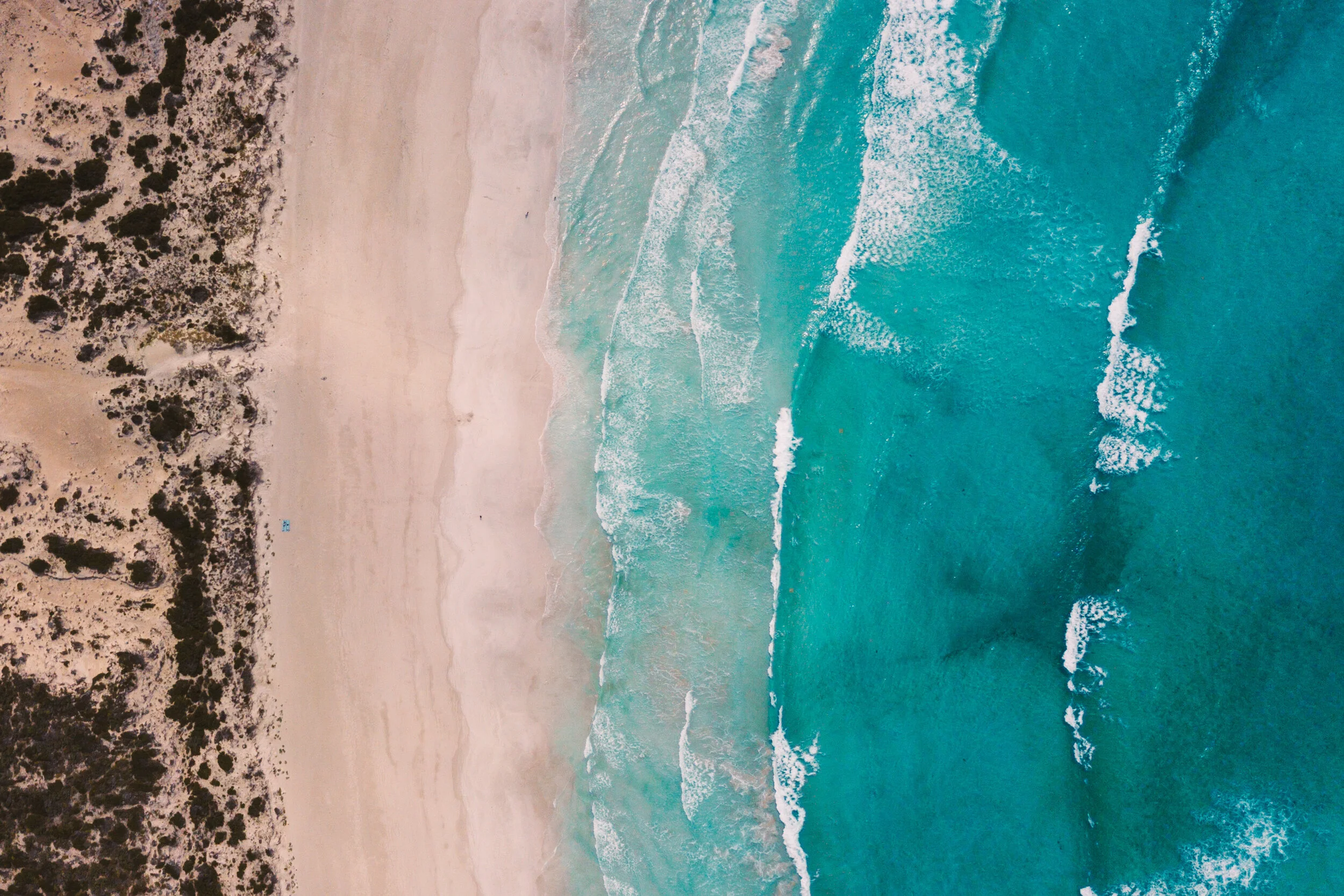 Aerial view of a sandy beach with waves crashing onto the shore, bordered by vegetation and dunes.