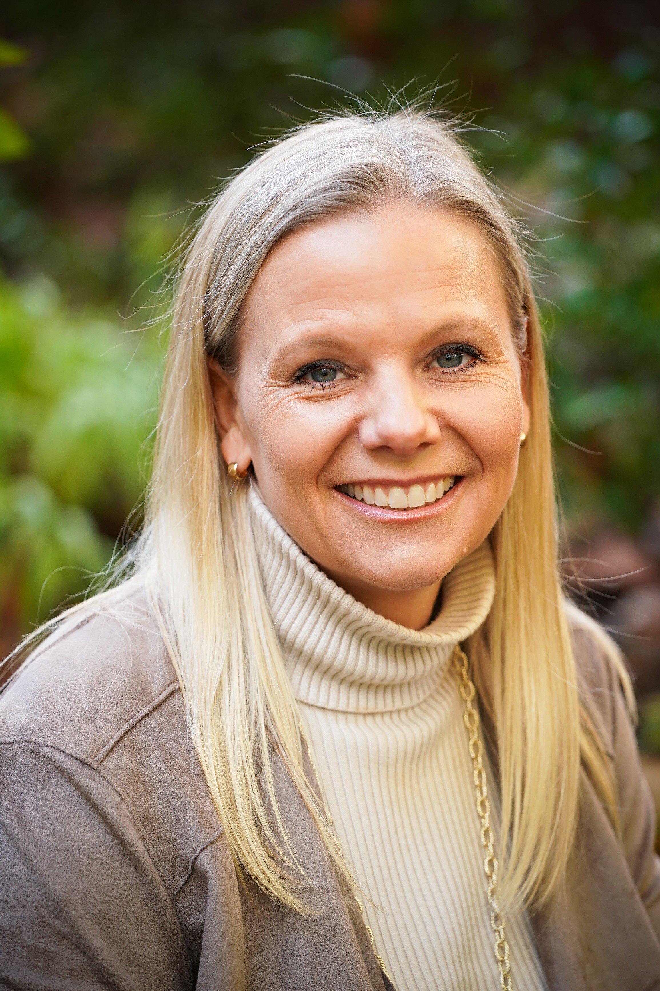 A smiling woman with blonde hair wearing earrings, a beige turtleneck sweater, and a brown coat, sitting outdoors with green blurred foliage in the background.