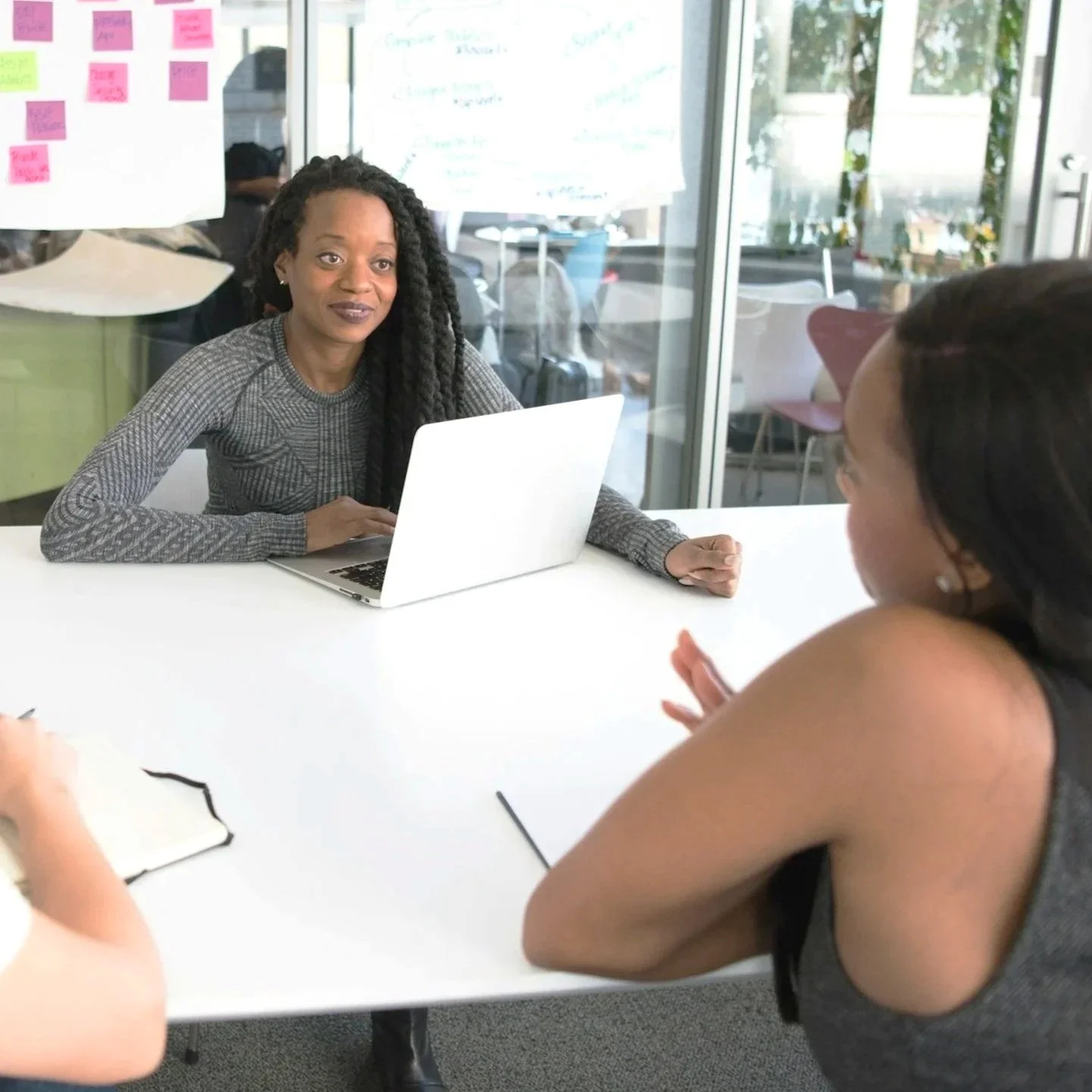 Three women are sitting around a white table during a meeting in a modern office. One woman with long black hair is seen from the side, leaning forward with her arm on the table. The woman across from her has long, braided black hair and is wearing a gray patterned top, sitting at a laptop. In the background, there are glass walls, colorful chairs, and large whiteboards with notes and charts.