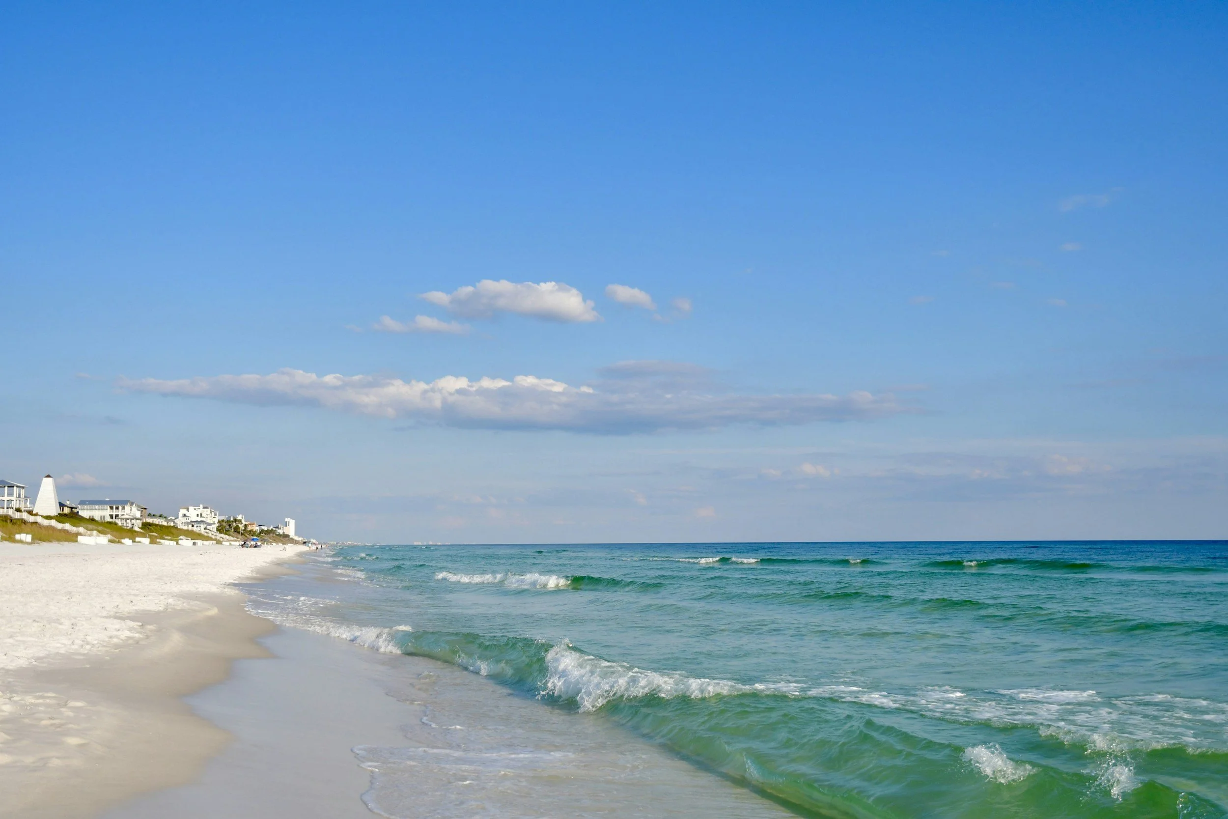 View of a beach with white sand and green waves, clear blue sky with some clouds, and beachfront houses in the distance.