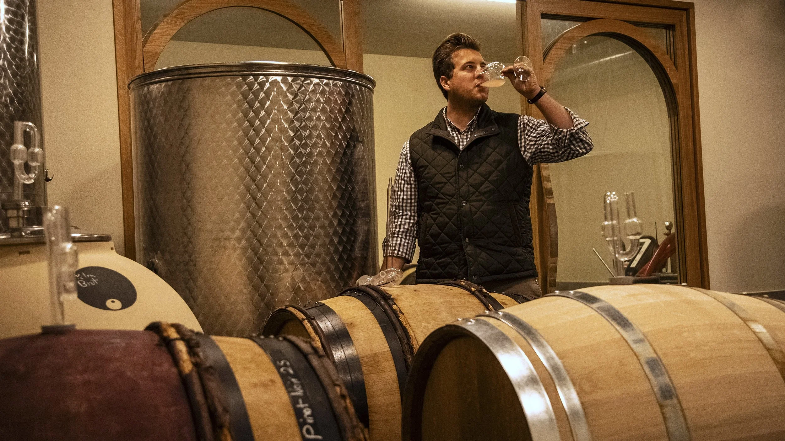 Ein junger Mann trinkt Wein aus einem Glas in einer Weinkellerei, umgeben von Holzfässern und einem großen Edelstahlbehälter.