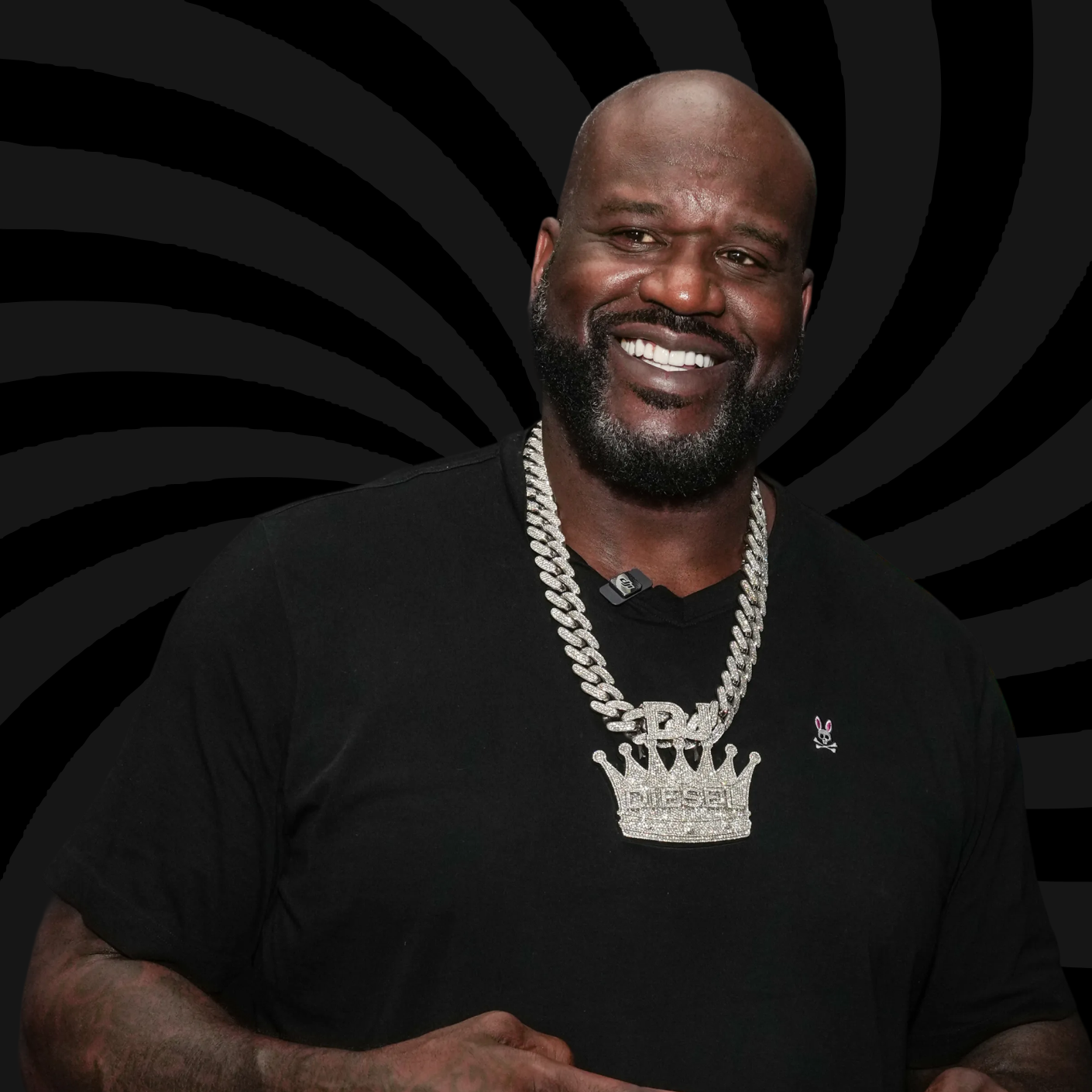 A smiling Black man with a beard, wearing a black shirt, a large silver chain with a crown pendant, and a small pink bunny pin, against a black background with spiral patterns.