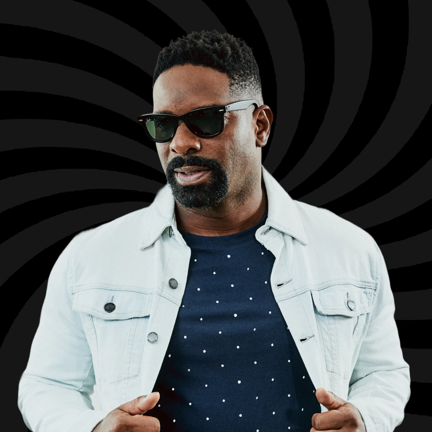 Man wearing sunglasses, a light-colored denim jacket, and a navy shirt with white polka dots, against a black background with spiral pattern.