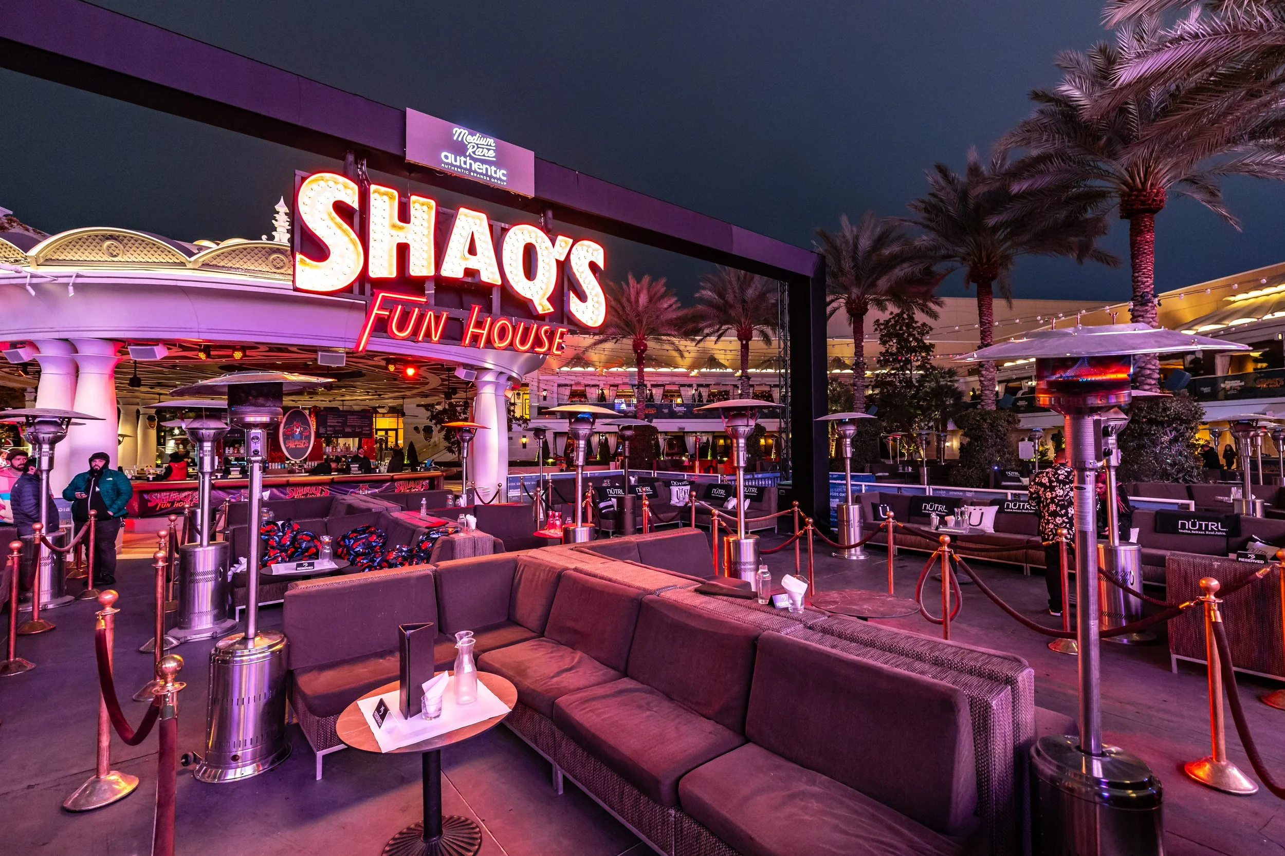 Outdoor seating area in front of Shaqs Fun House with purple and pink neon signs, palm trees, and patio heaters at night.