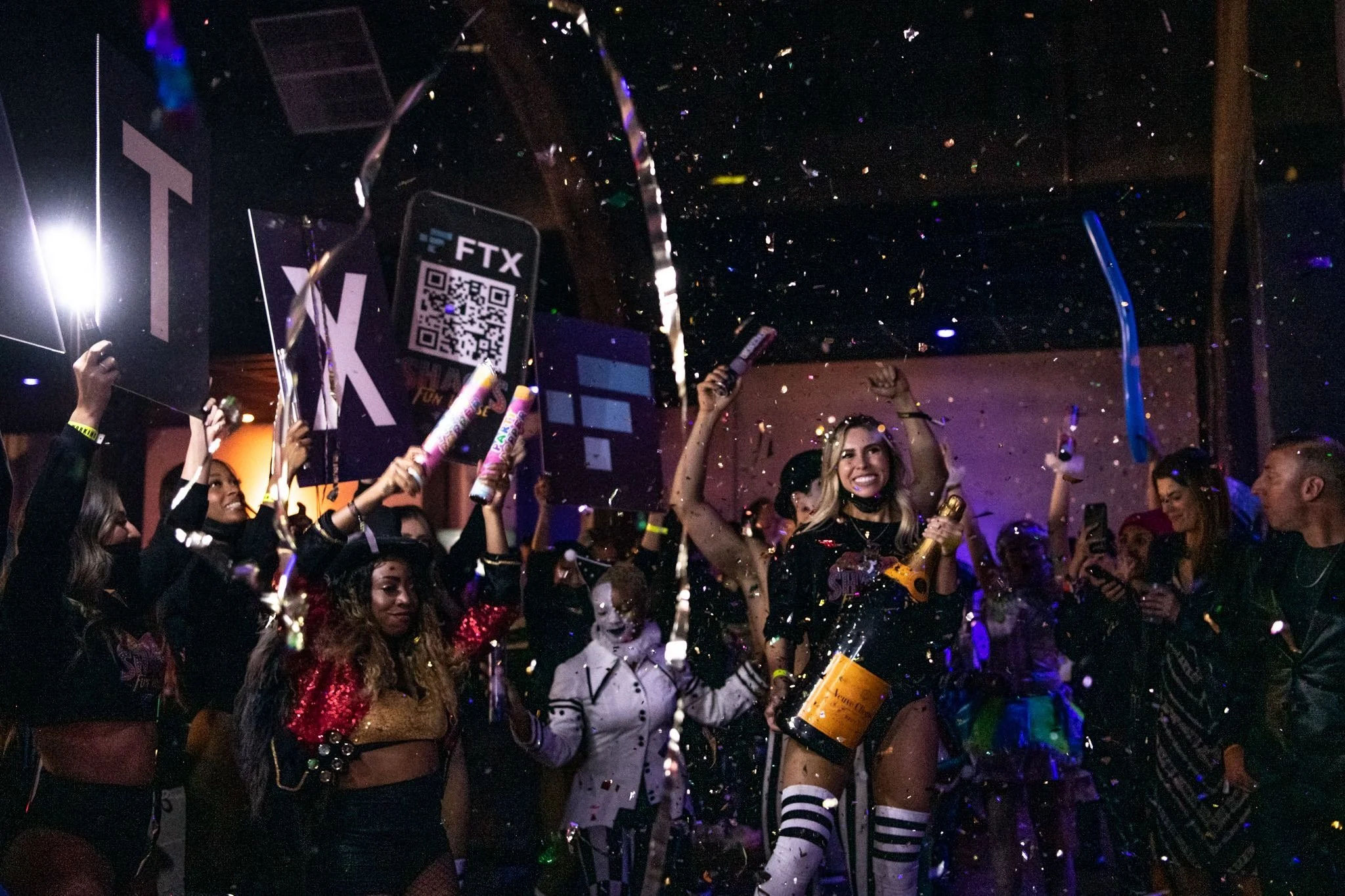 People celebrating with confetti, some holding signs, in a lively party setting, with one woman holding a large champagne bottle and smiling.