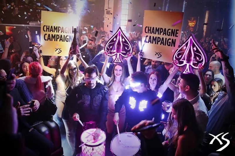 Crowd celebrating at a party with signs that read 'Champagne Campaign' and playing drums, with freestyle dance and confetti, in a vibrant nightclub setting.