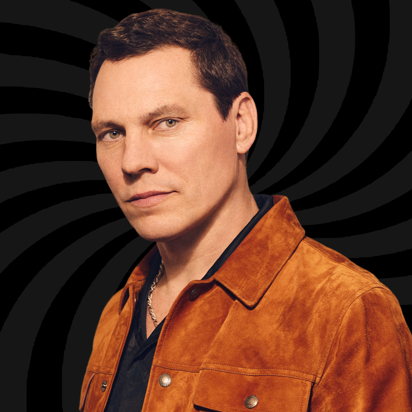 A man with short dark hair wearing a brown leather jacket over a black shirt with a chain necklace, posing against a black background with a spiral pattern.