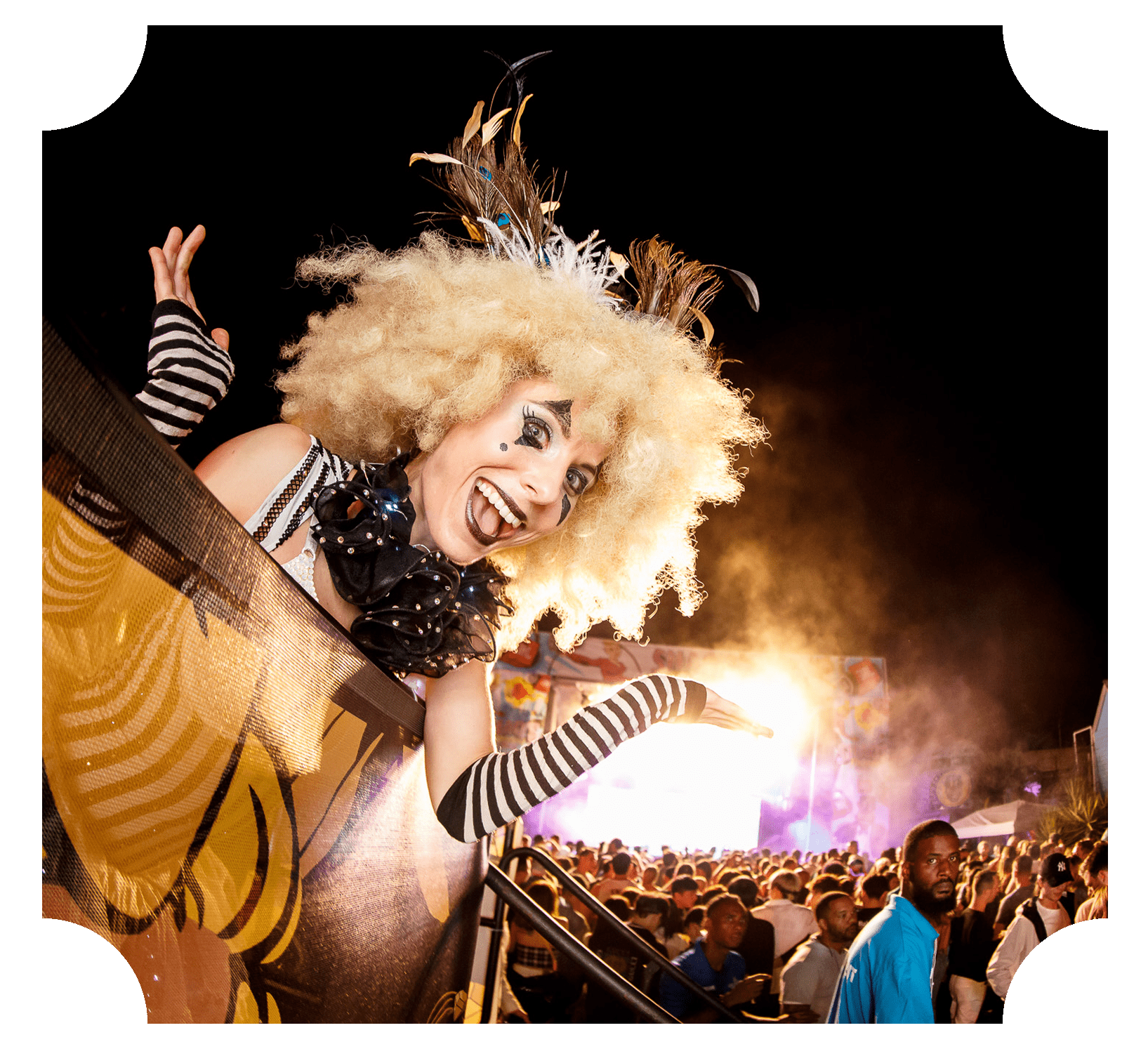 A woman in a costume with a big blonde curly wig, feathered headpiece, clown makeup, and striped gloves at a concert or festival, with a large crowd and stage with lights in the background.