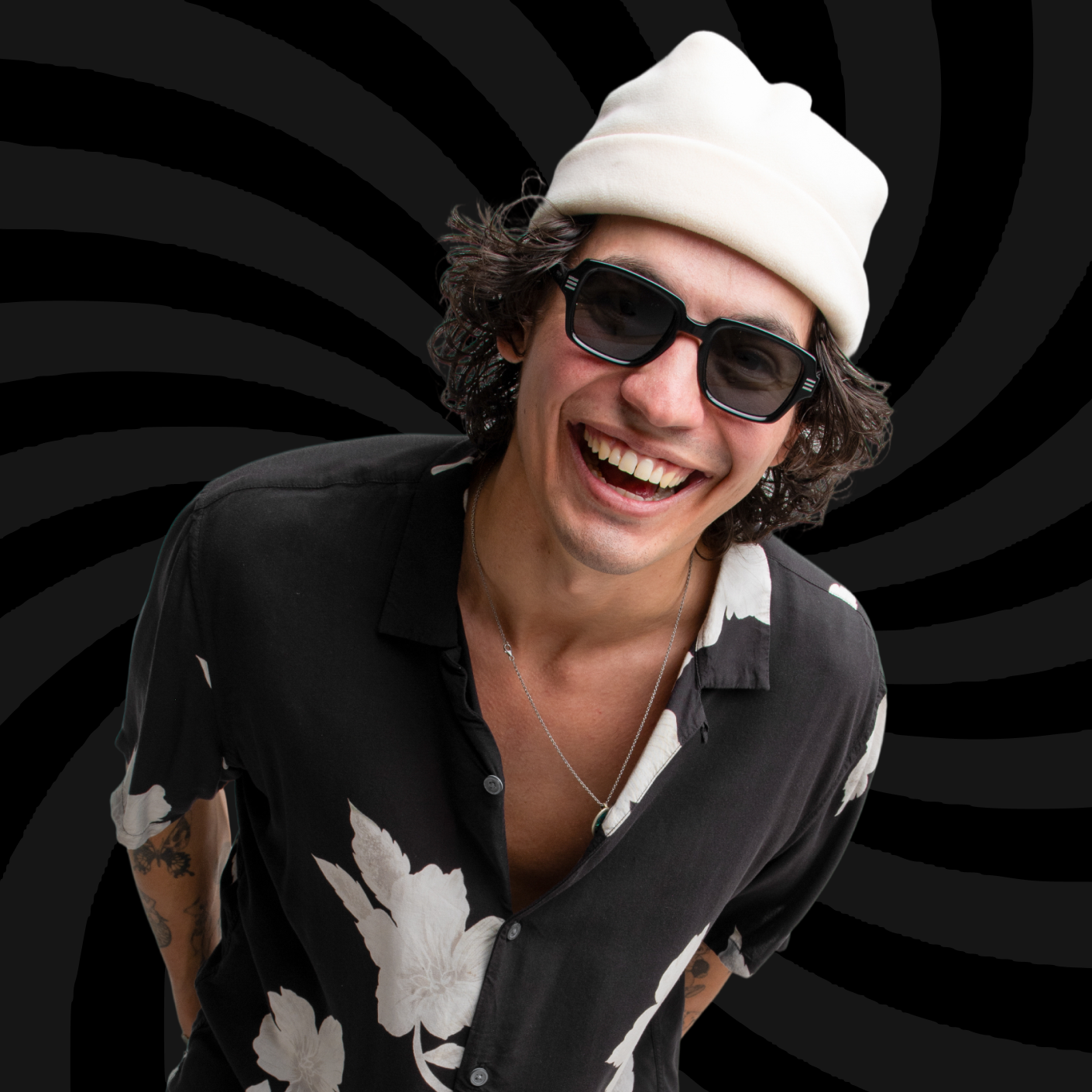 Smiling young man with curly hair, wearing a white beanie, sunglasses, a black and white floral shirt, and a necklace, against a black spiral background.
