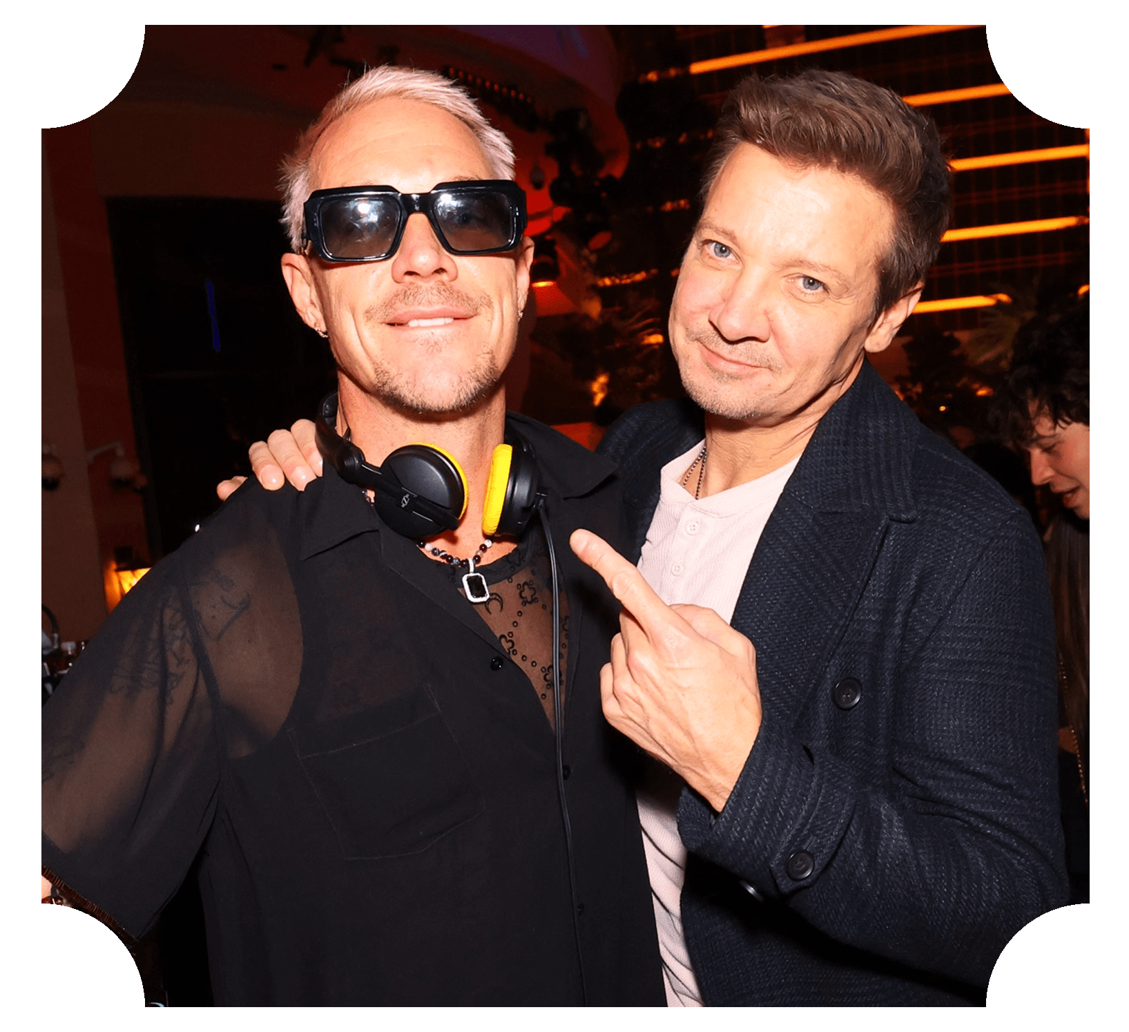Two men posing together at a party; one wearing sunglasses, headphones around his neck, and a black shirt; the other wearing a dark jacket and pointing at the first man.