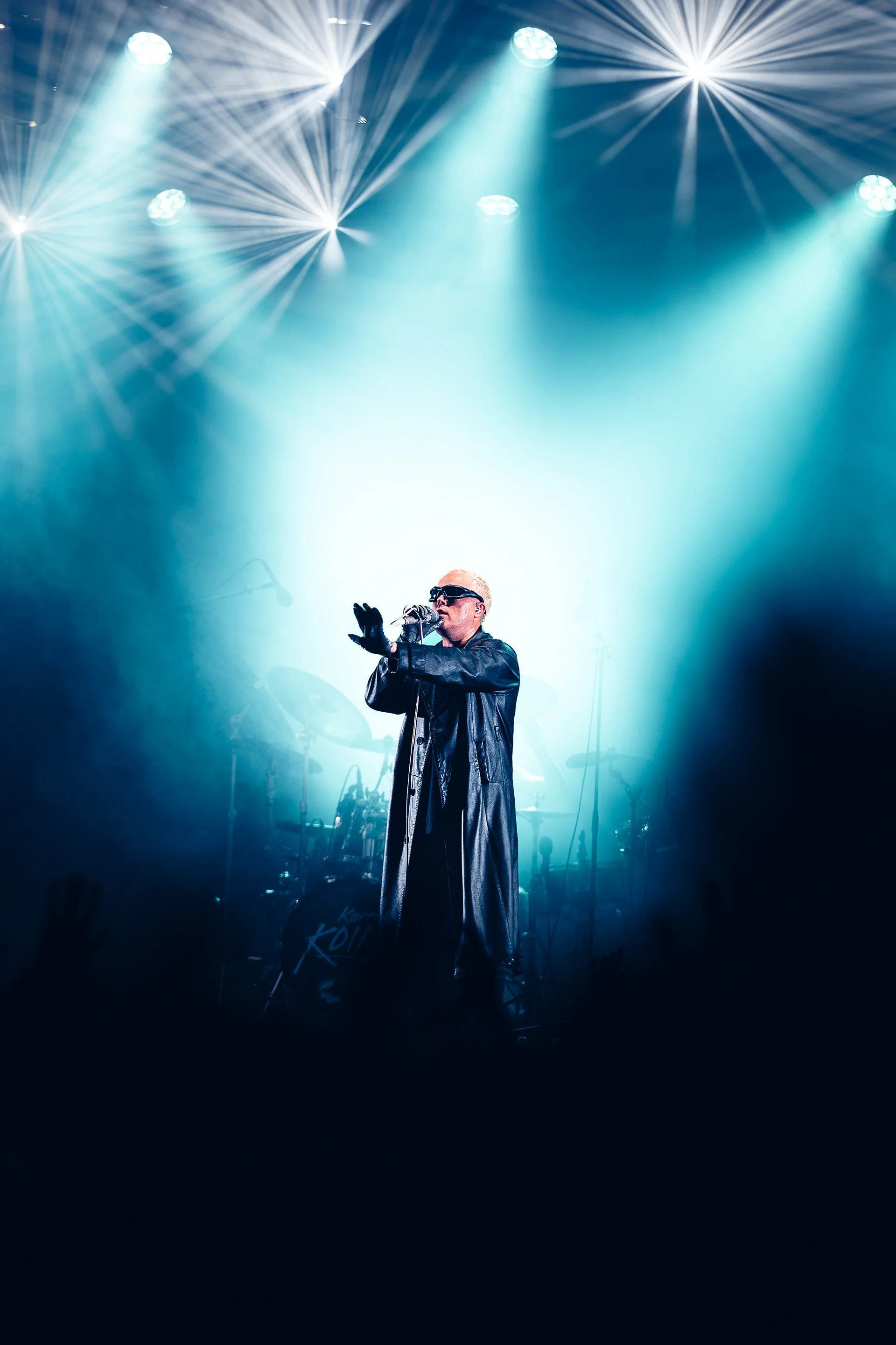 Performer on stage singing into a microphone with bright stage lights shining down, wearing sunglasses and a long black leather coat.