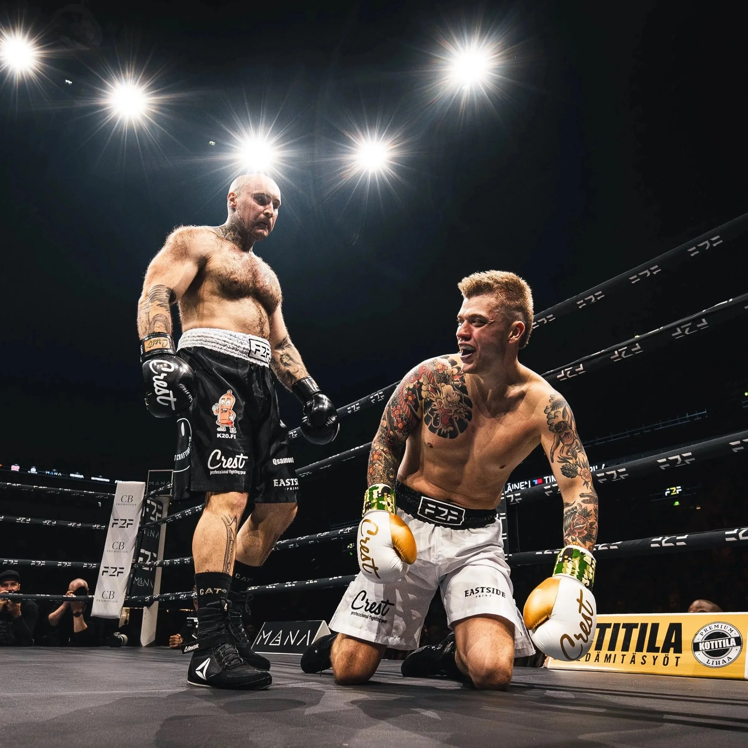 Two mixed martial arts fighters in a ring after a match. One fighter is standing, the other is kneeling and smiling. The standing fighter appears disappointed.