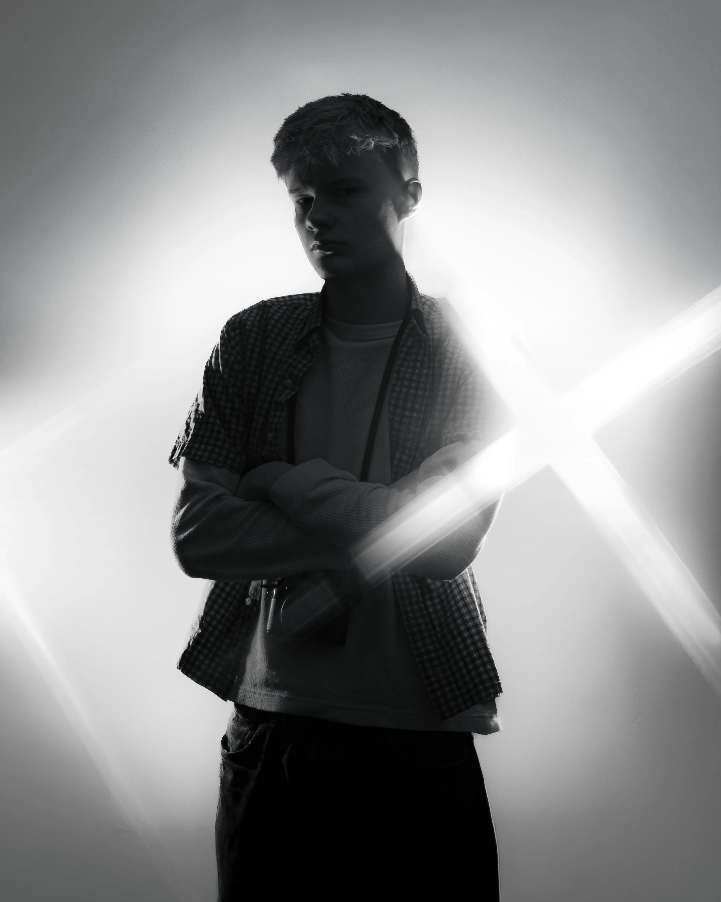 Black and white photo of a young man with crossed arms, wearing a checkered shirt over a plain t-shirt, standing with backlit lighting creating a silhouette effect.