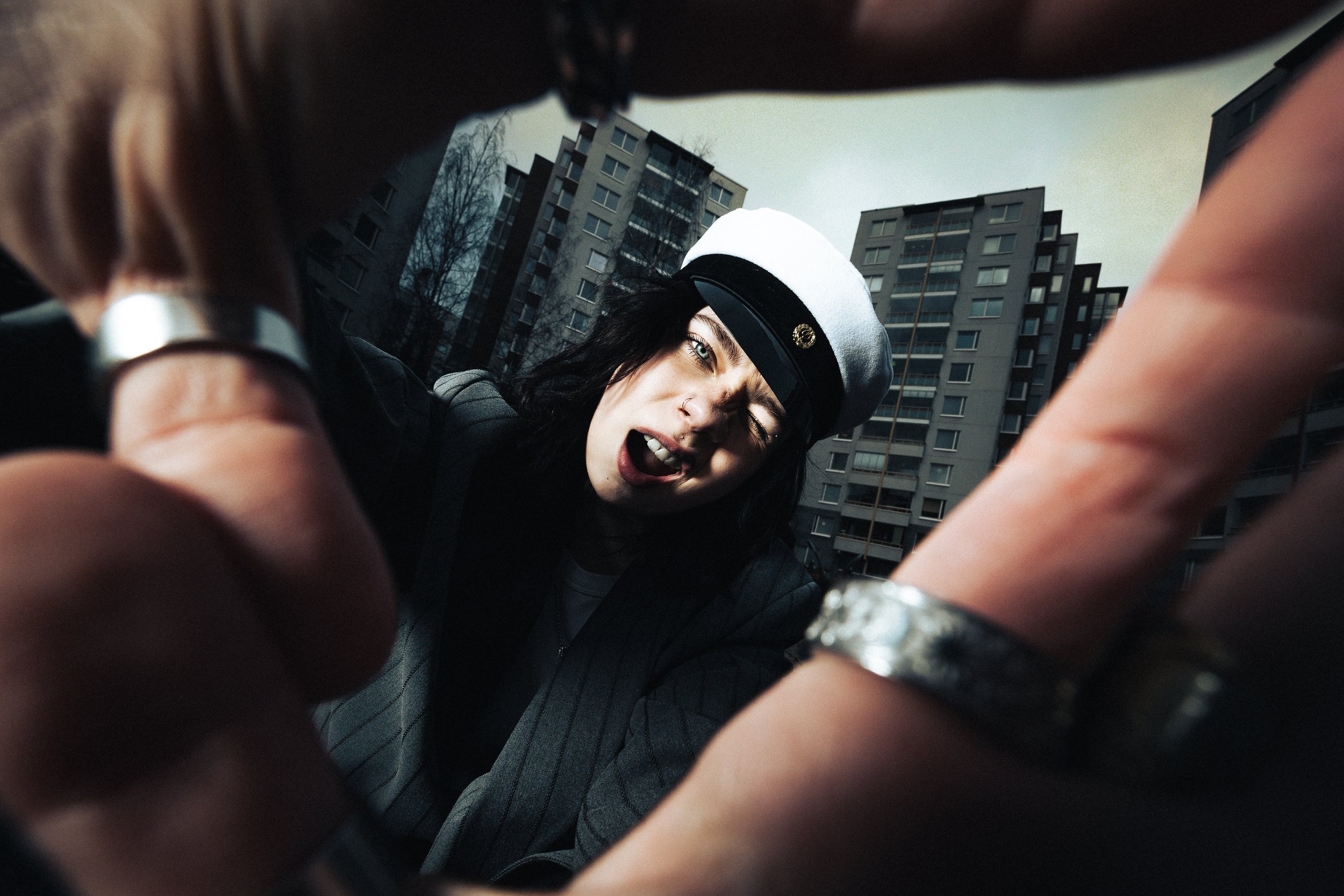 Woman with black hair wearing a white hat with a black band, making an angry or shouting expression, with urban buildings in the background.