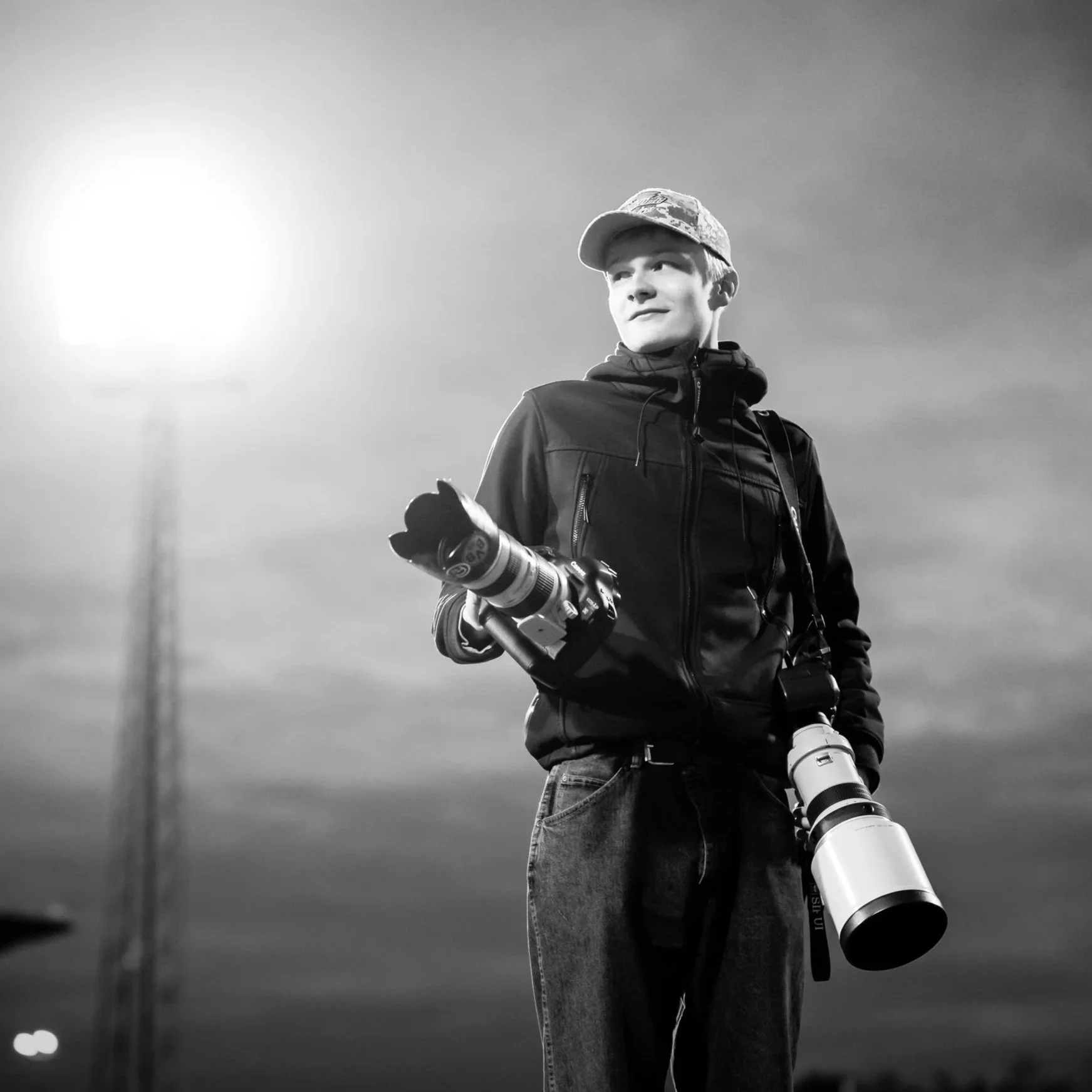 A young man outdoors at dusk holding a camera with a telephoto lens, wearing a jacket and cap, with a blurred background of the sky and a tall structure.
