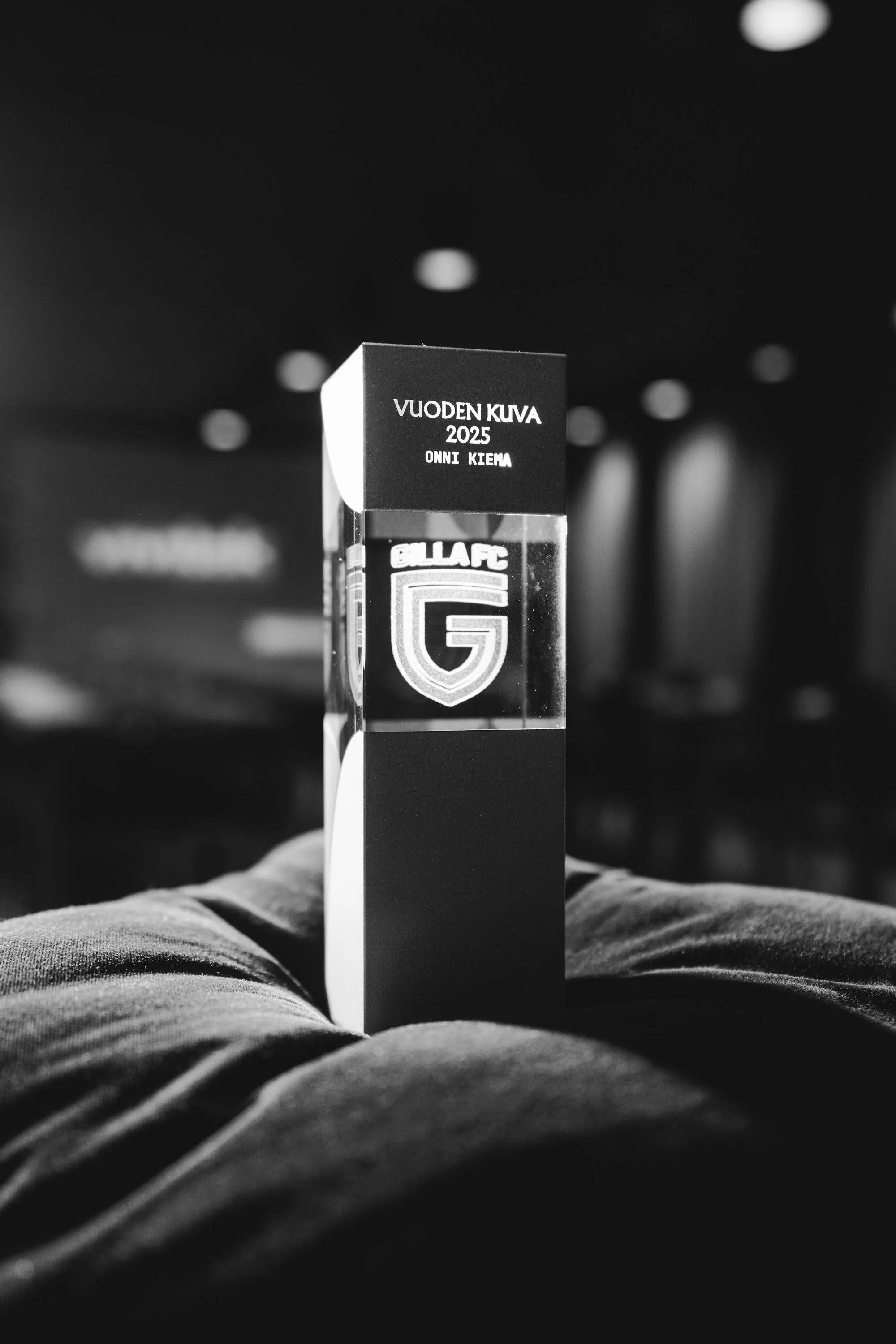 Black and white photo of a rectangular, transparent trophy with the logo 'G' and the text 'BULLAFC' on it, resting on a person's pants in a dimly lit room.