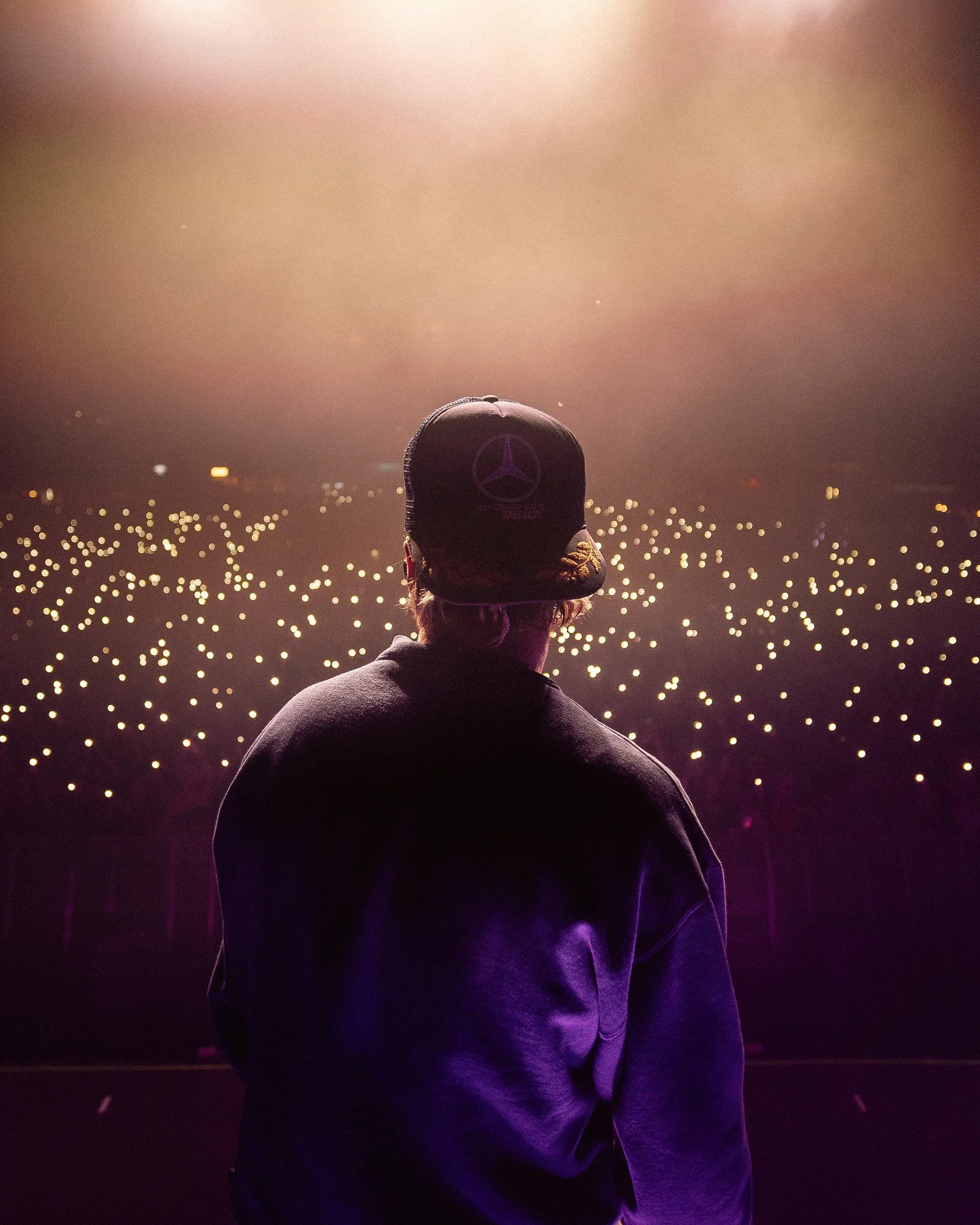 Performer on stage facing an audience with lights, wearing a cap with a Mercedes-Benz logo.