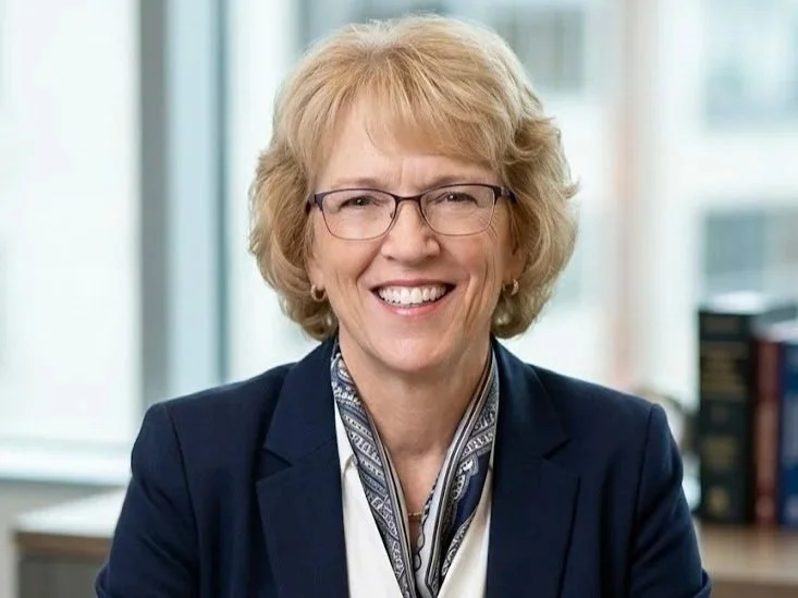 A smiling middle-aged woman with blonde hair, glasses, and a dark blazer sitting in an office with large windows.