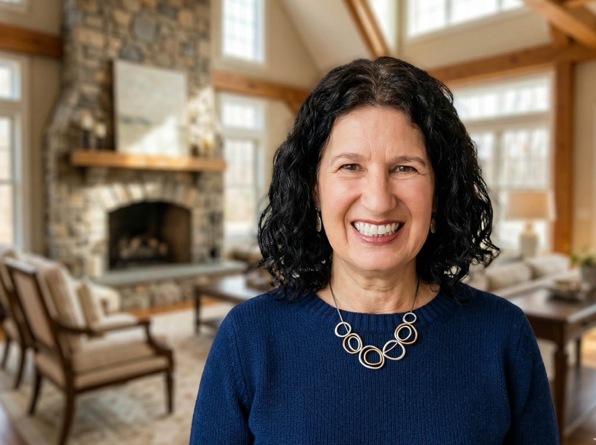 A smiling woman with dark curly hair wearing a blue sweater and a circular silver necklace in a cozy living room with a stone fireplace, large windows, and wooden accents.