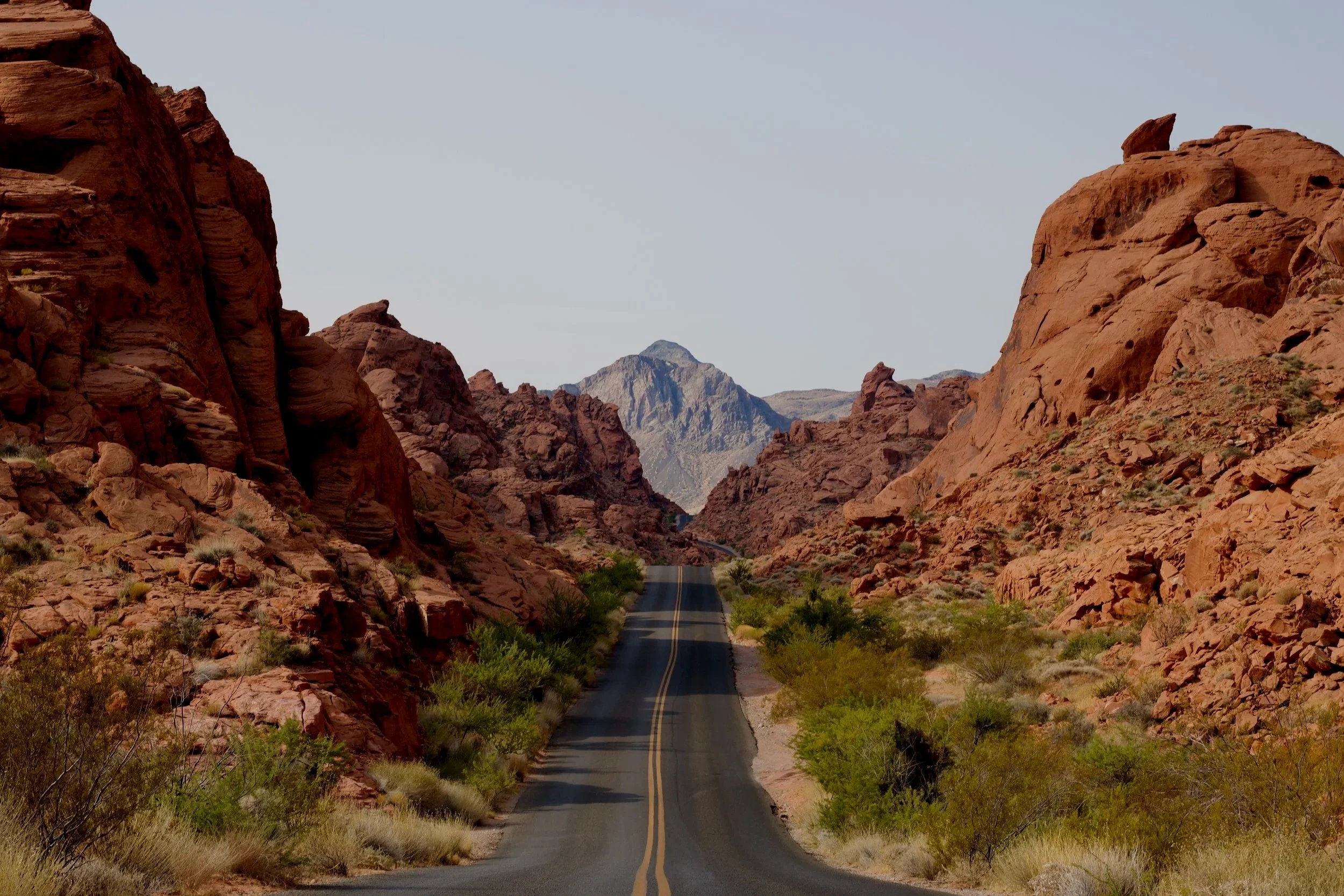 A two-lane road winding through a desert canyon with red rock formations on either side and a mountain in the distance under a clear sky.