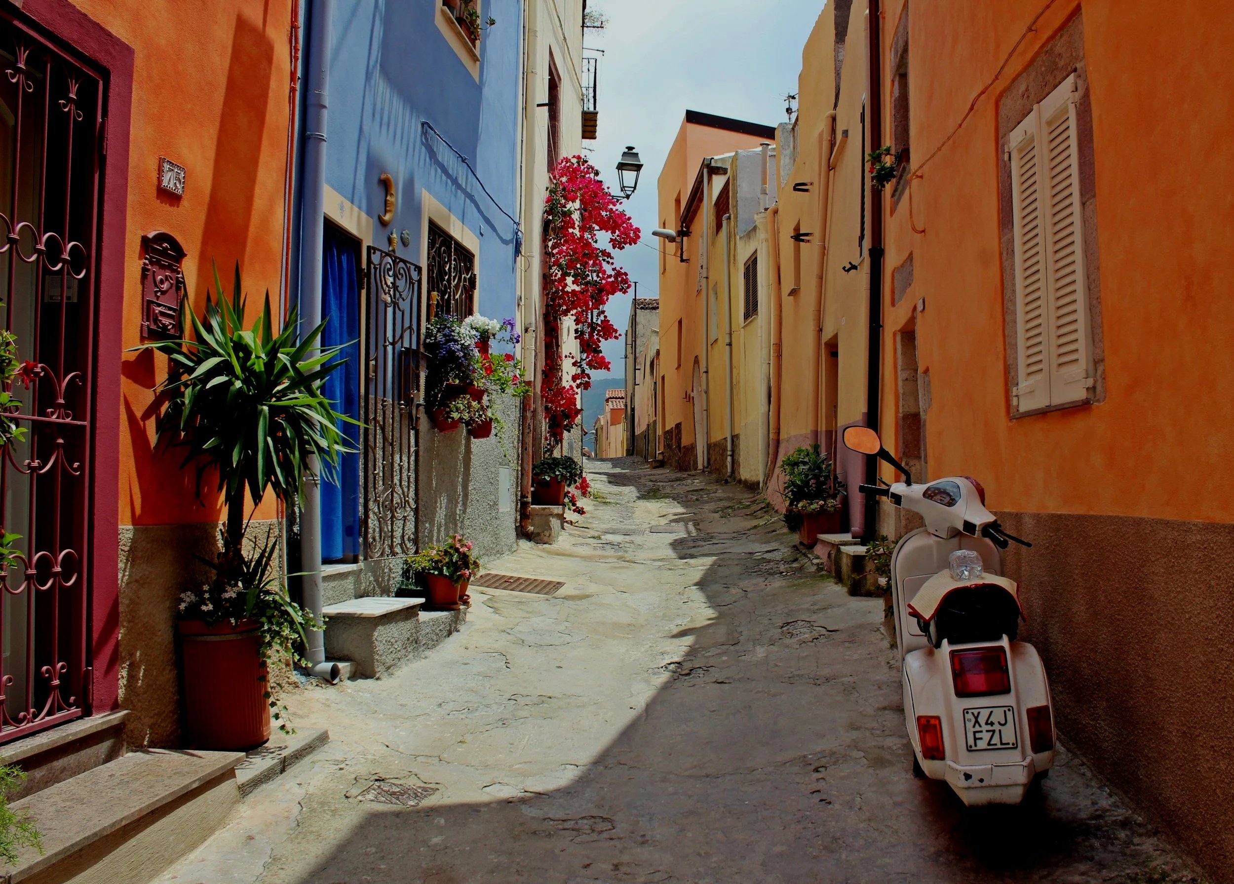 Colorful narrow street with pastel-colored houses, potted plants, and a white scooter parked on the side, in a Mediterranean village.