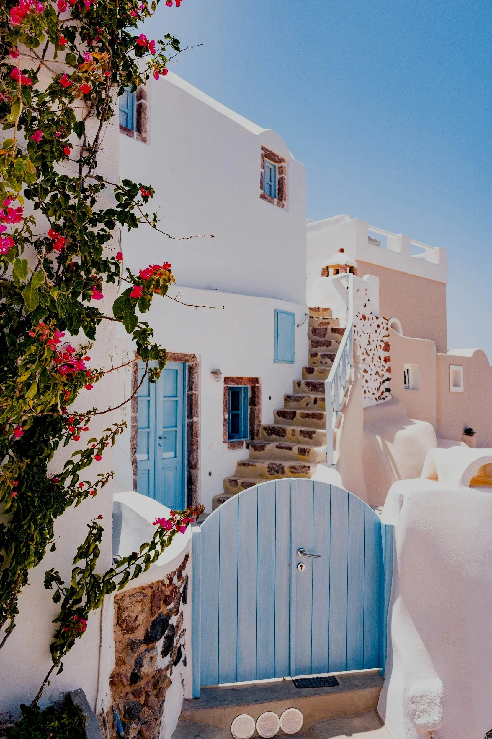 White Mediterranean-style building with blue doors and windows, stone staircase, and pink flowering vines against a bright blue sky.