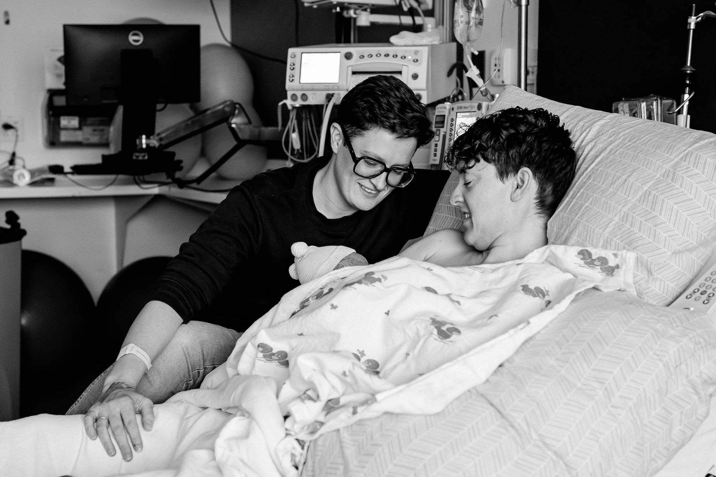A woman with glasses and a short hairstyle is smiling and leaning over a bed, holding hands with a young man who is lying in the hospital bed, both appearing happy.