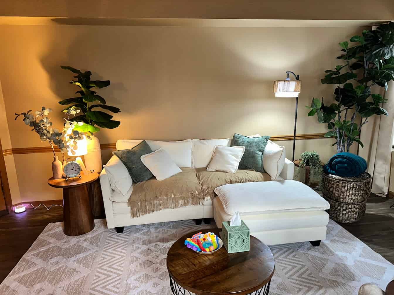 Living room with a white sofa, cream throw blanket, and multiple pillows, surrounded by green plants, a wooden side table with lamp and clock, a round coffee table, and a basket with blanket, under warm lighting.