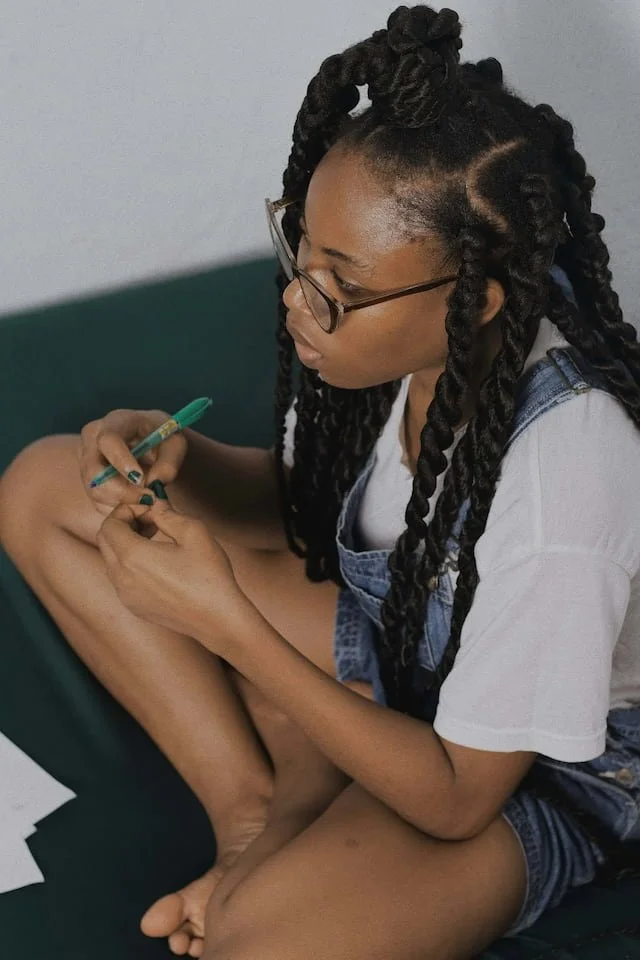 A young woman with braids and glasses sitting cross-legged, writing with a pen on a piece of paper. A woman after EMDR therapy.