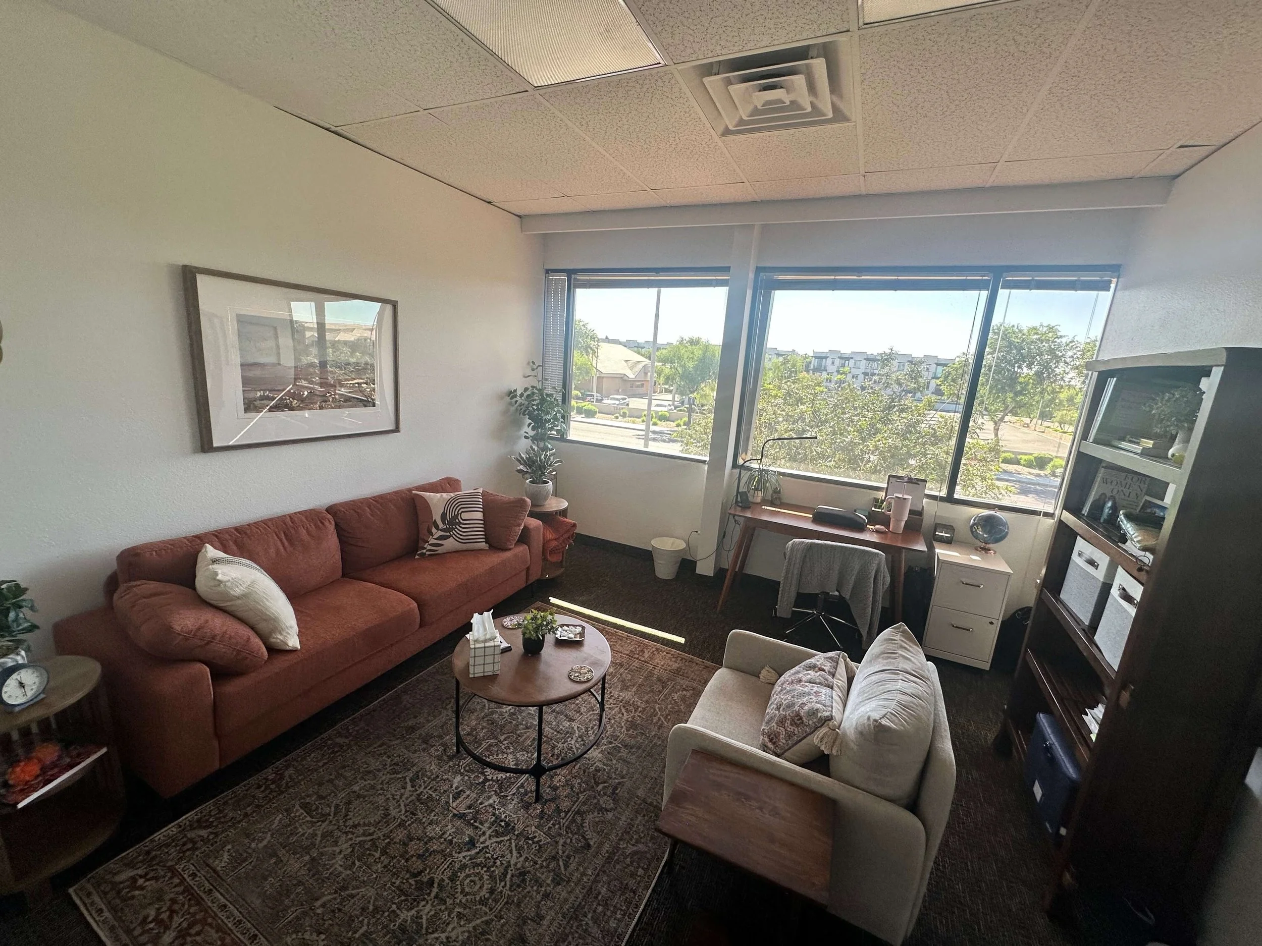 A cozy trauma therapist's office with a large window. Contains a pink sofa with pillows, a small round coffee table, a white armchair with a pillow, a wooden desk with a chair, and a dark bookshelf. 