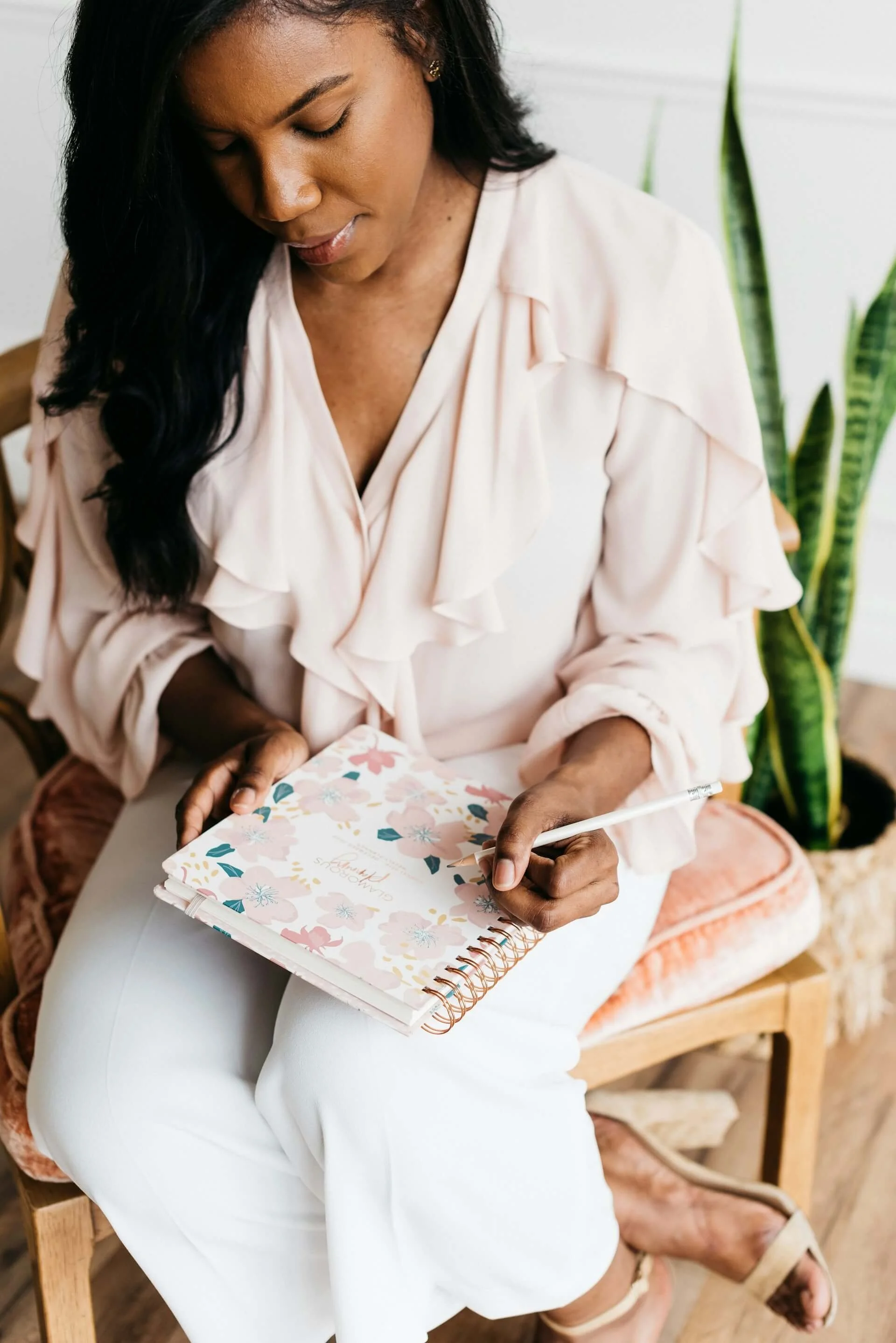 Woman writing in a floral-patterned planner seated on a pink cushion chair with a green plant in the background reflecting on what she learned in therapy